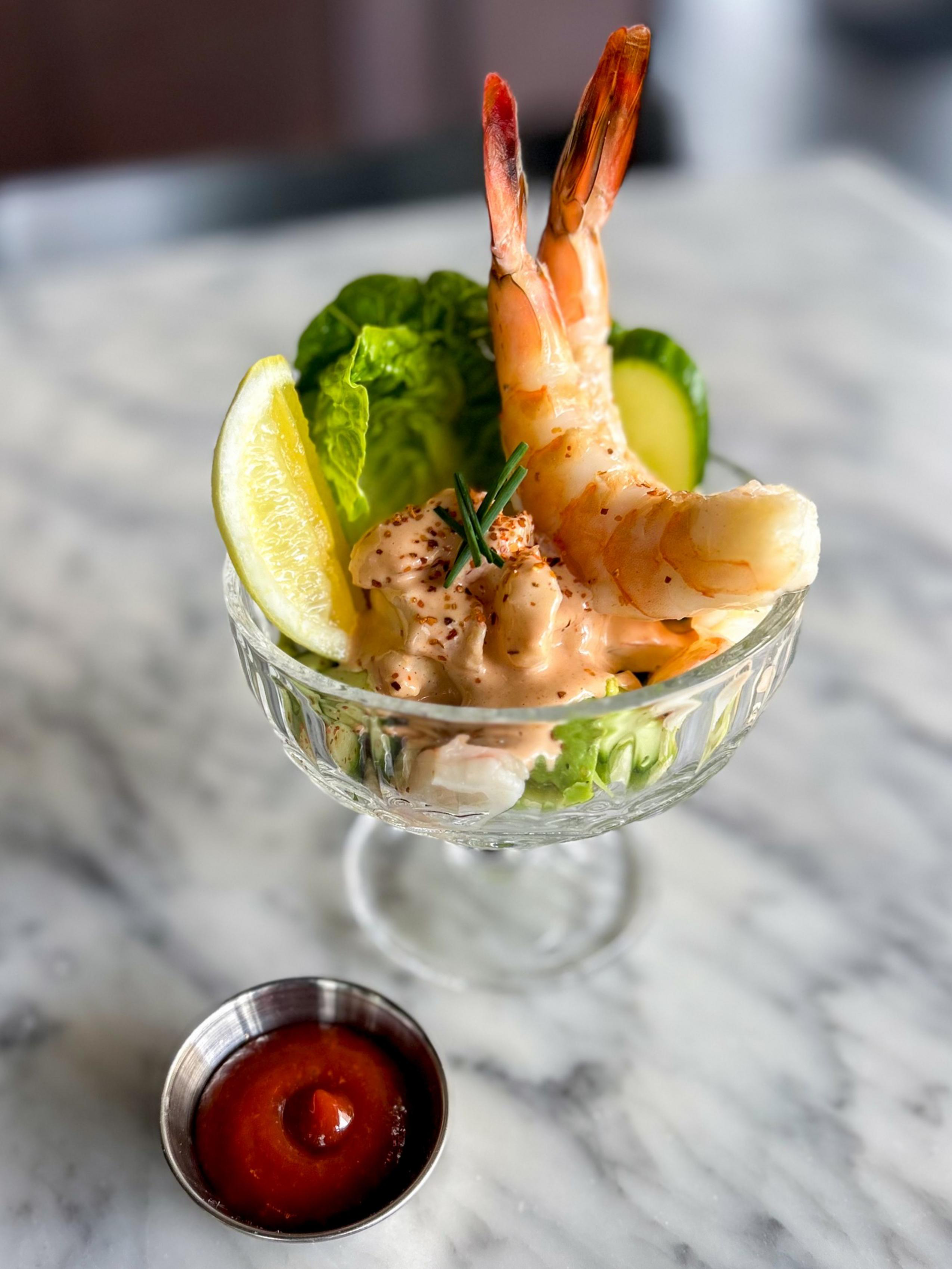 A glass dish holds cocktail shrimp, lettuce, cucumber slice, lemon wedge, and creamy sauce, with a small metal cup of cocktail sauce beside it.