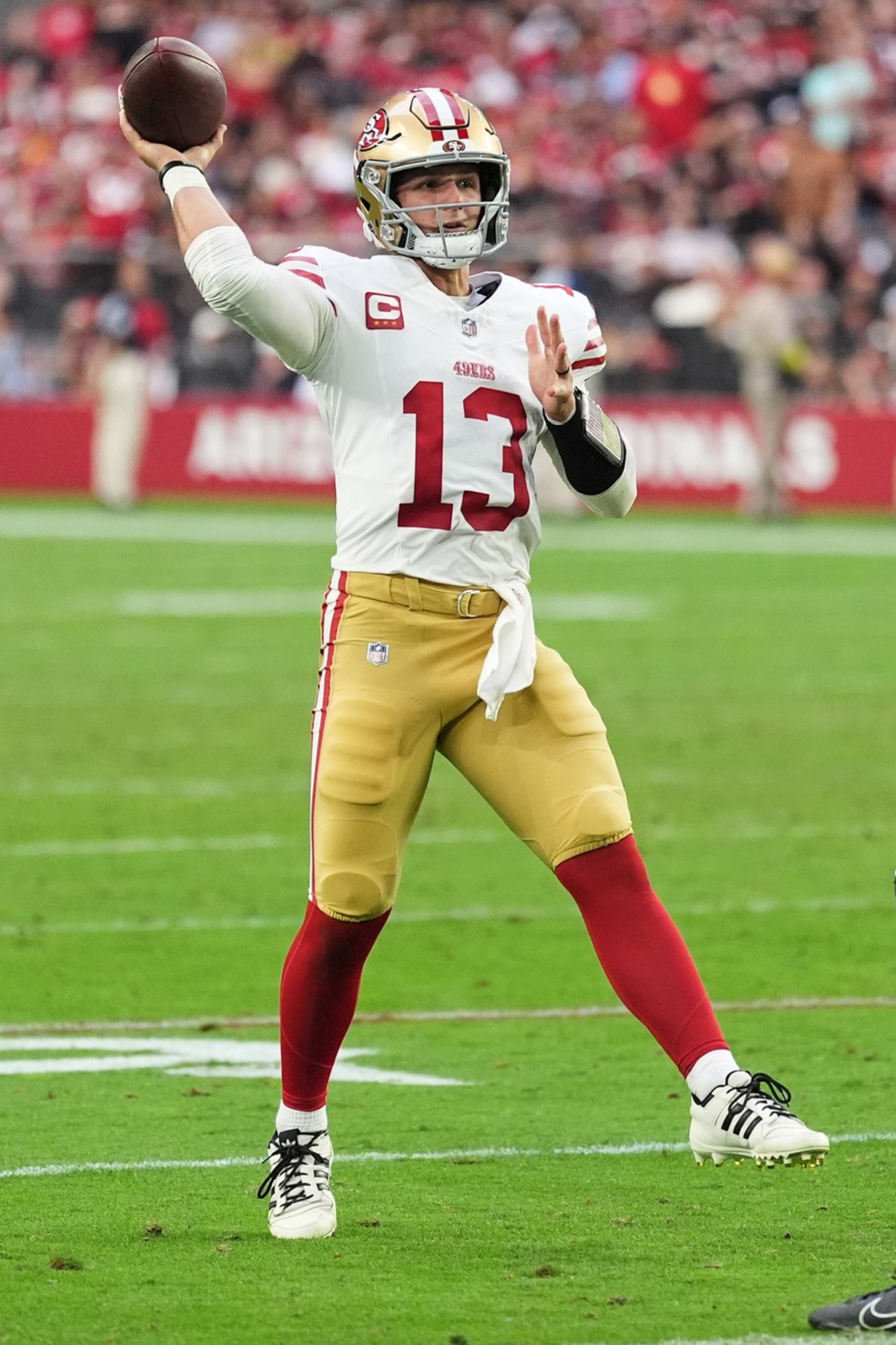A football player in white and gold prepares to throw a pass while a player in black with number 16 approaches defensively on the field.