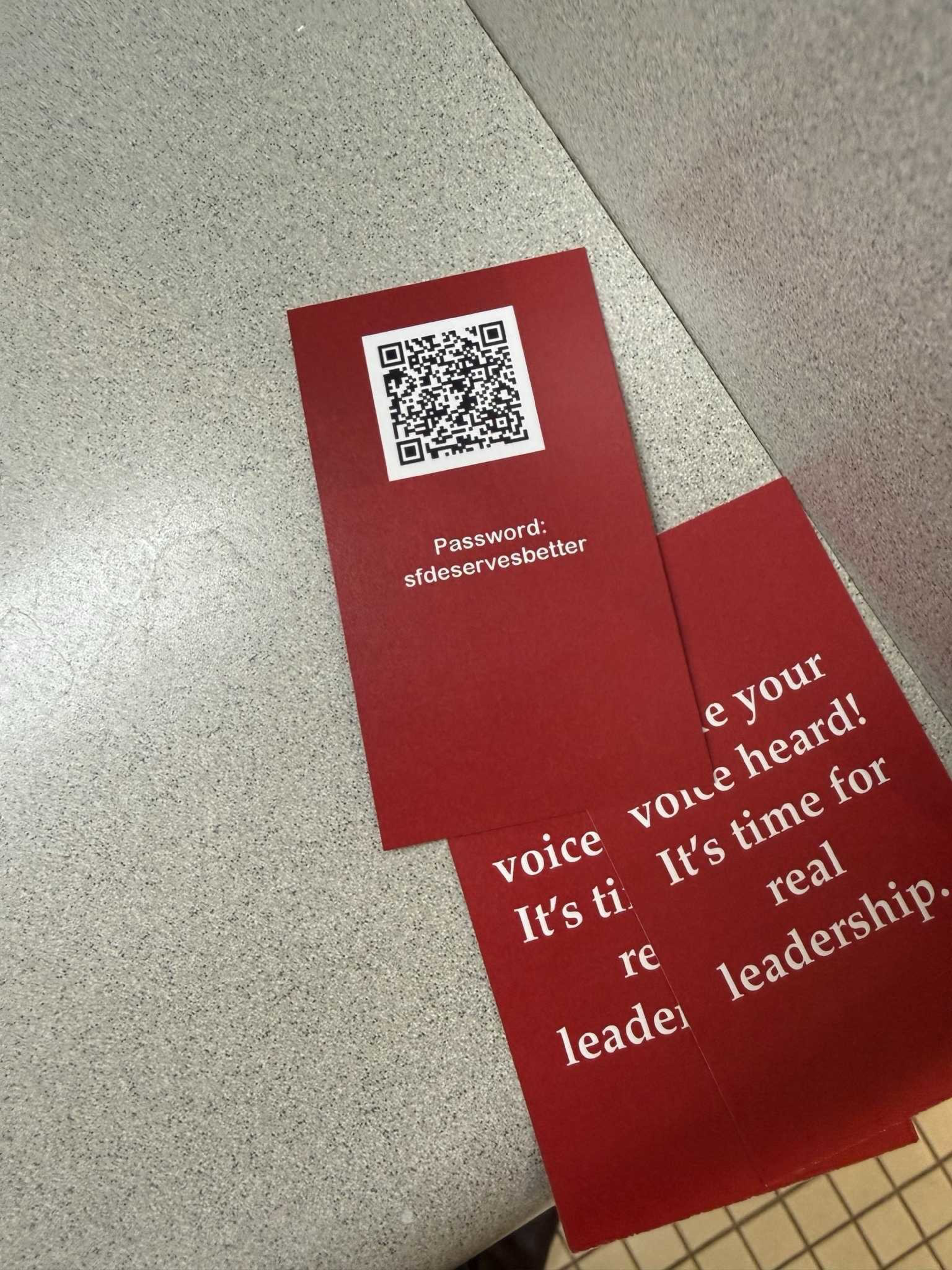 A red card with a QR code and the password “sfdeservesbetter” lies on a gray countertop, partially covering another red card with white text about leadership.