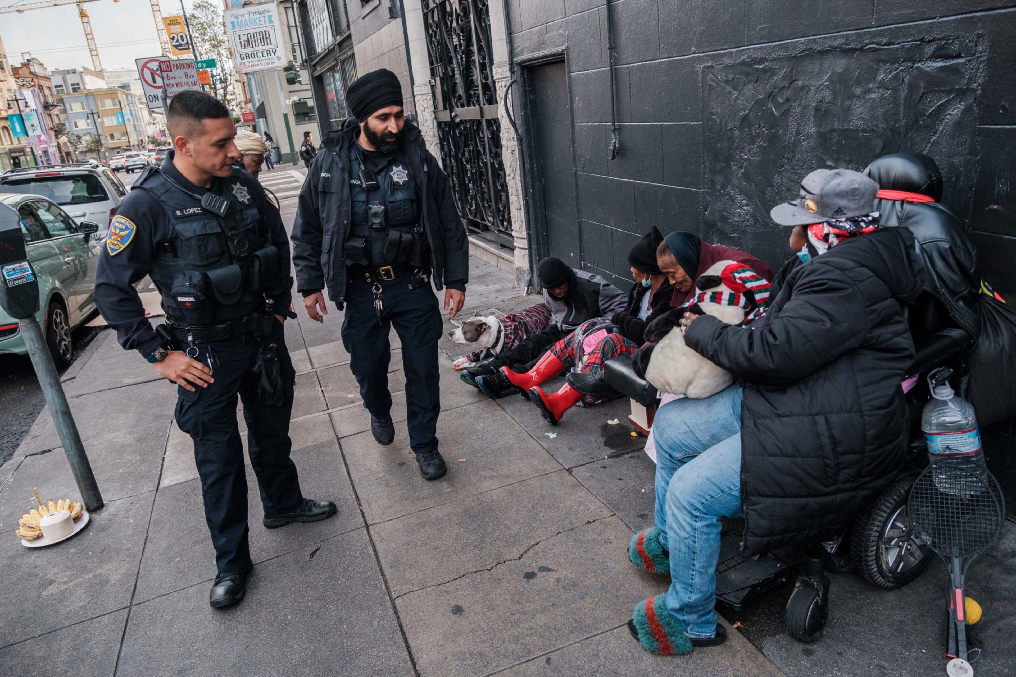 Two police officers stand on a sidewalk near a group of seated individuals, some wearing colorful clothing, next to a black wall and a parked scooter.
