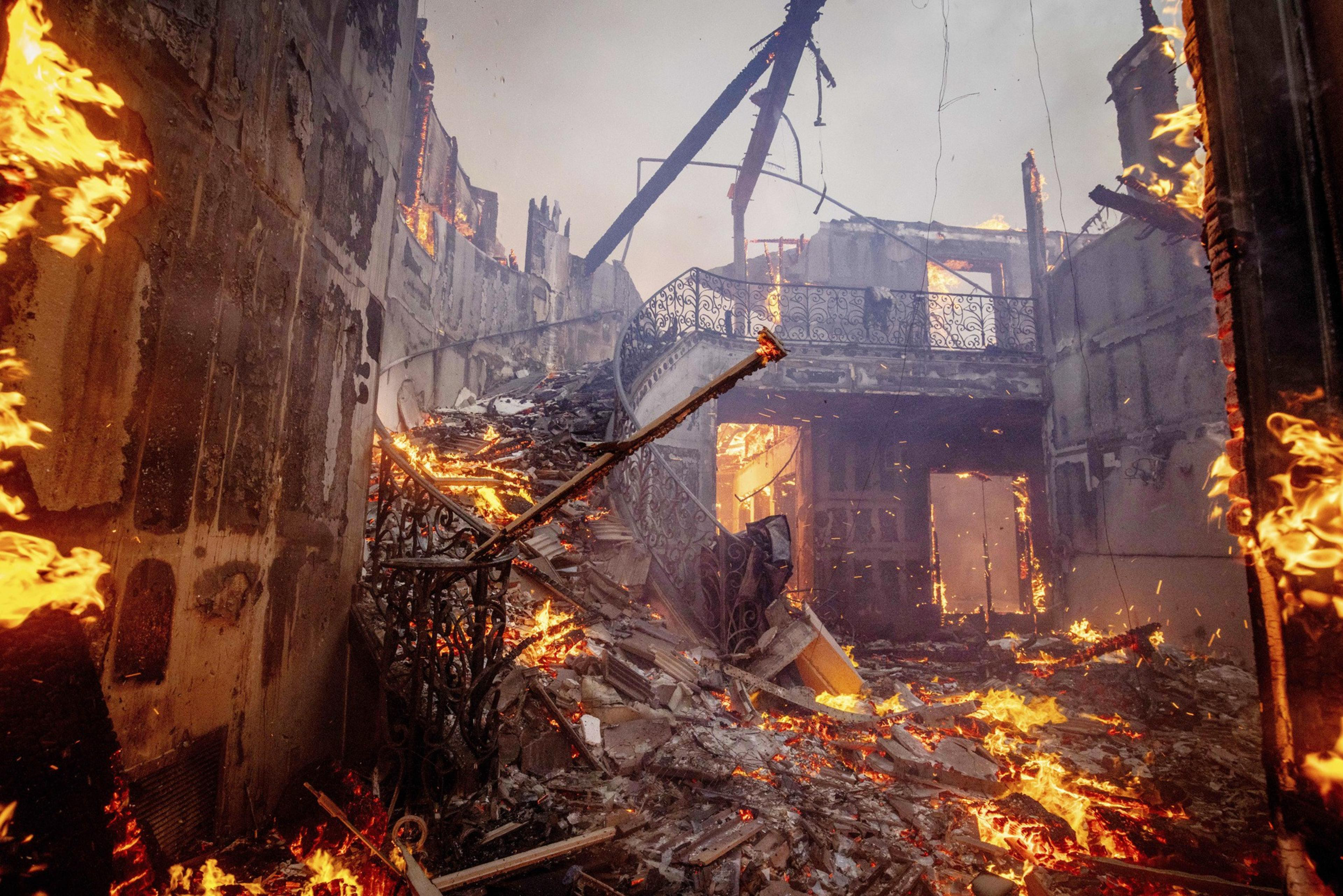 A large, destroyed indoor structure is engulfed in flames, with debris scattered across the floor and charred remains of walls and railings visible.