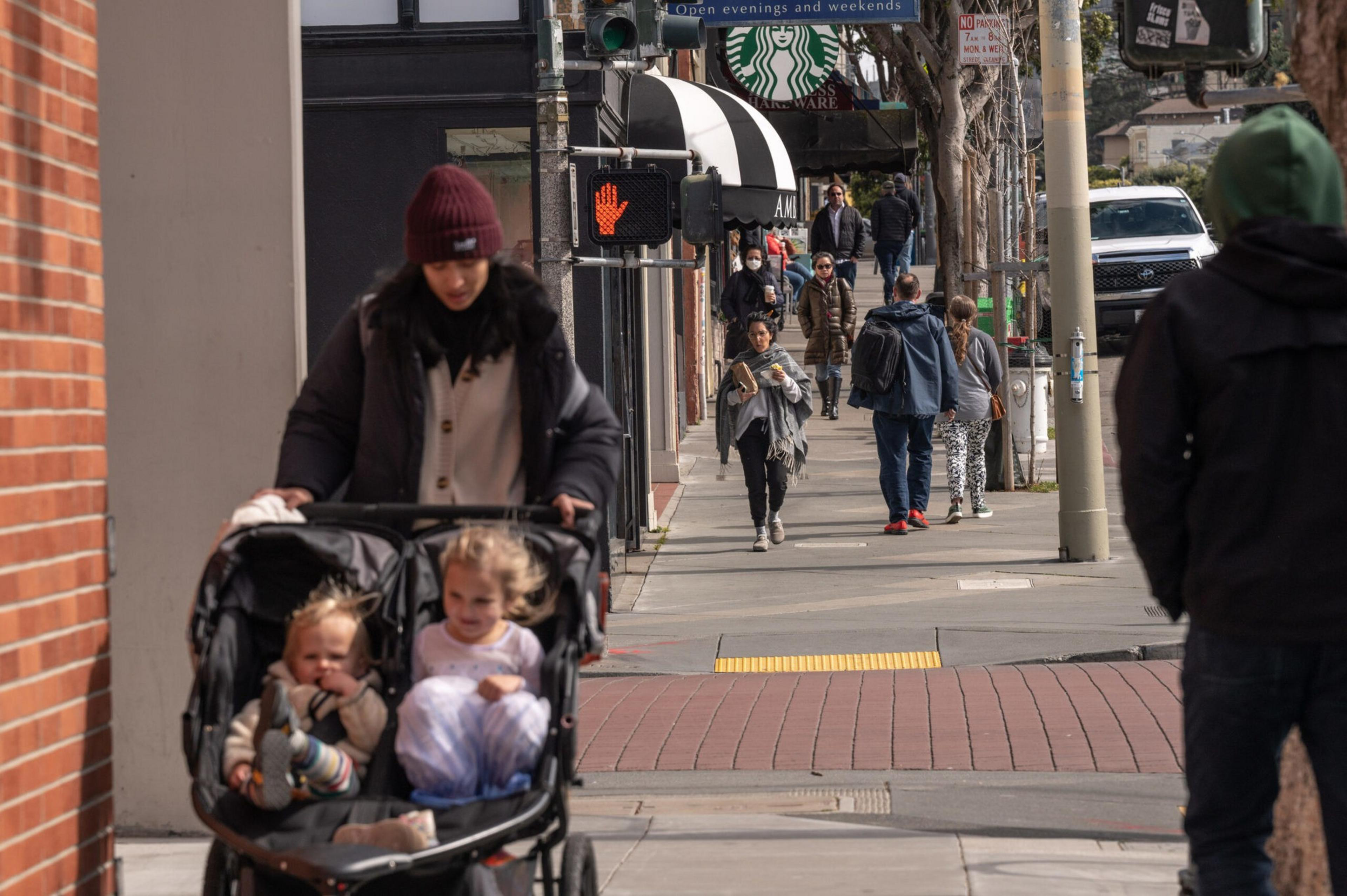 A woman pushes a double stroller with two young children along a city sidewalk while several pedestrians walk in the background near a Starbucks.