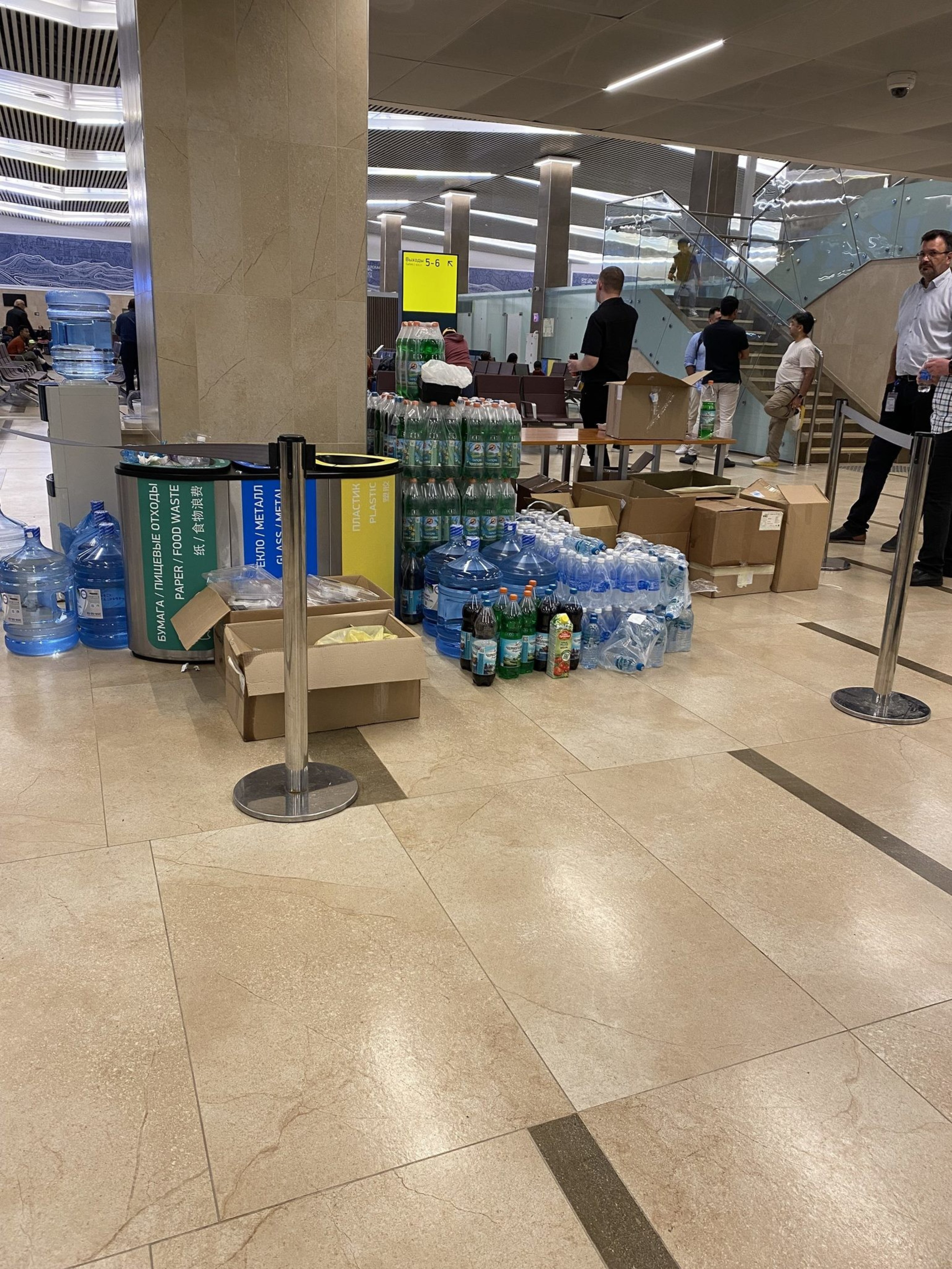 The image shows a modern indoor public space with people. It features stacks of bottled water and cleaning supplies, surrounded by various waste bins and some open boxes.