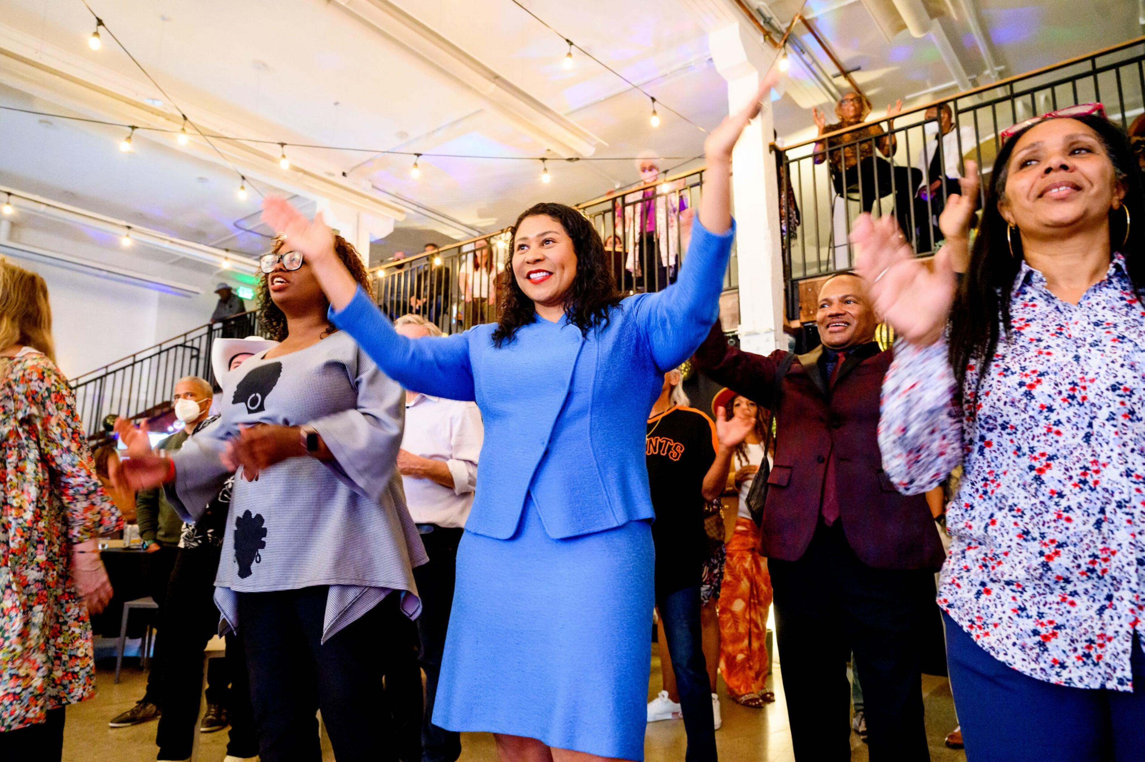 A group of diverse people indoors are smiling and clapping with raised hands, appearing joyful and engaged in a lively event or celebration.