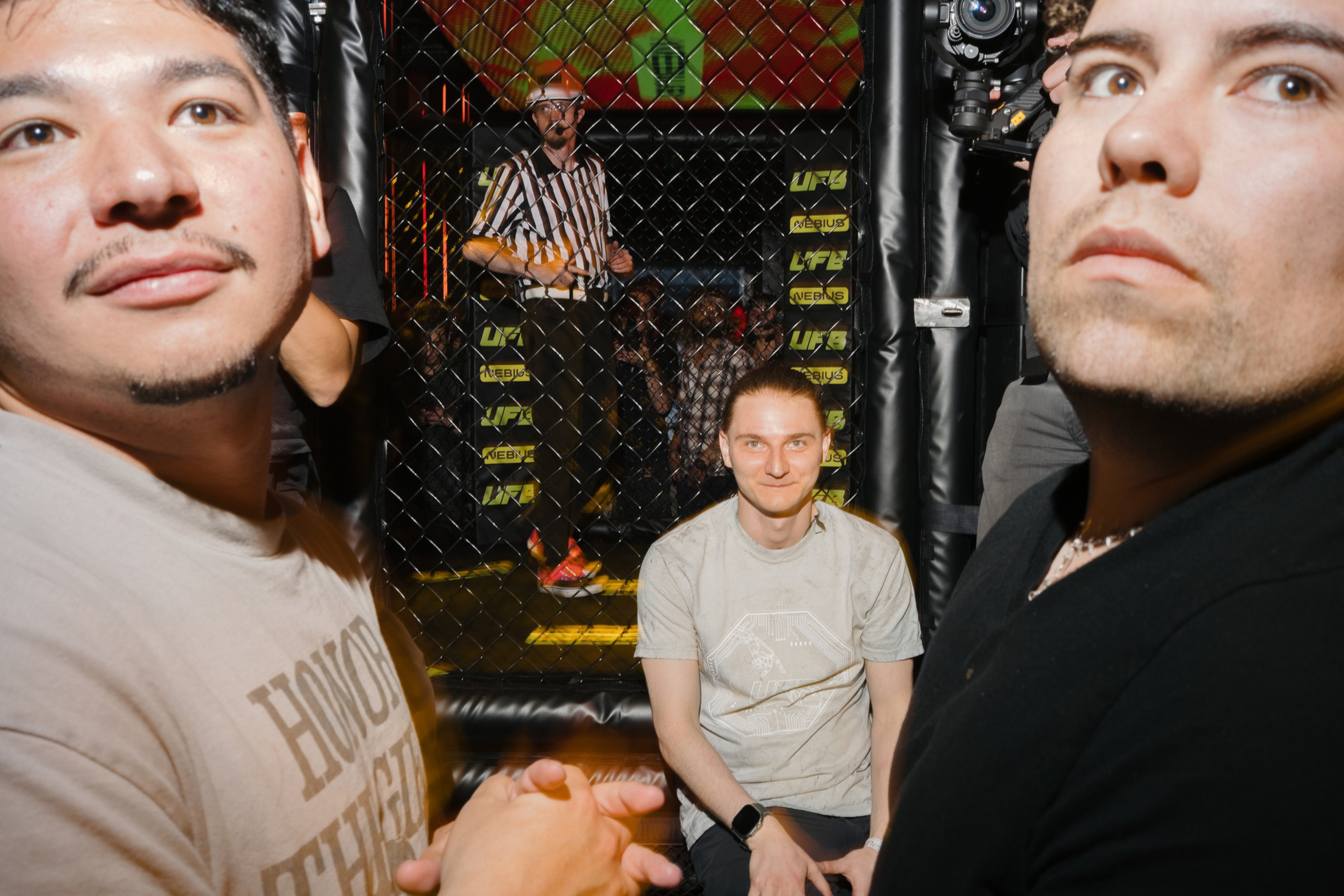 Two men stand close to the camera near an MMA cage, while a third person sits inside the cage and a referee stands in the background behind the cage fence.