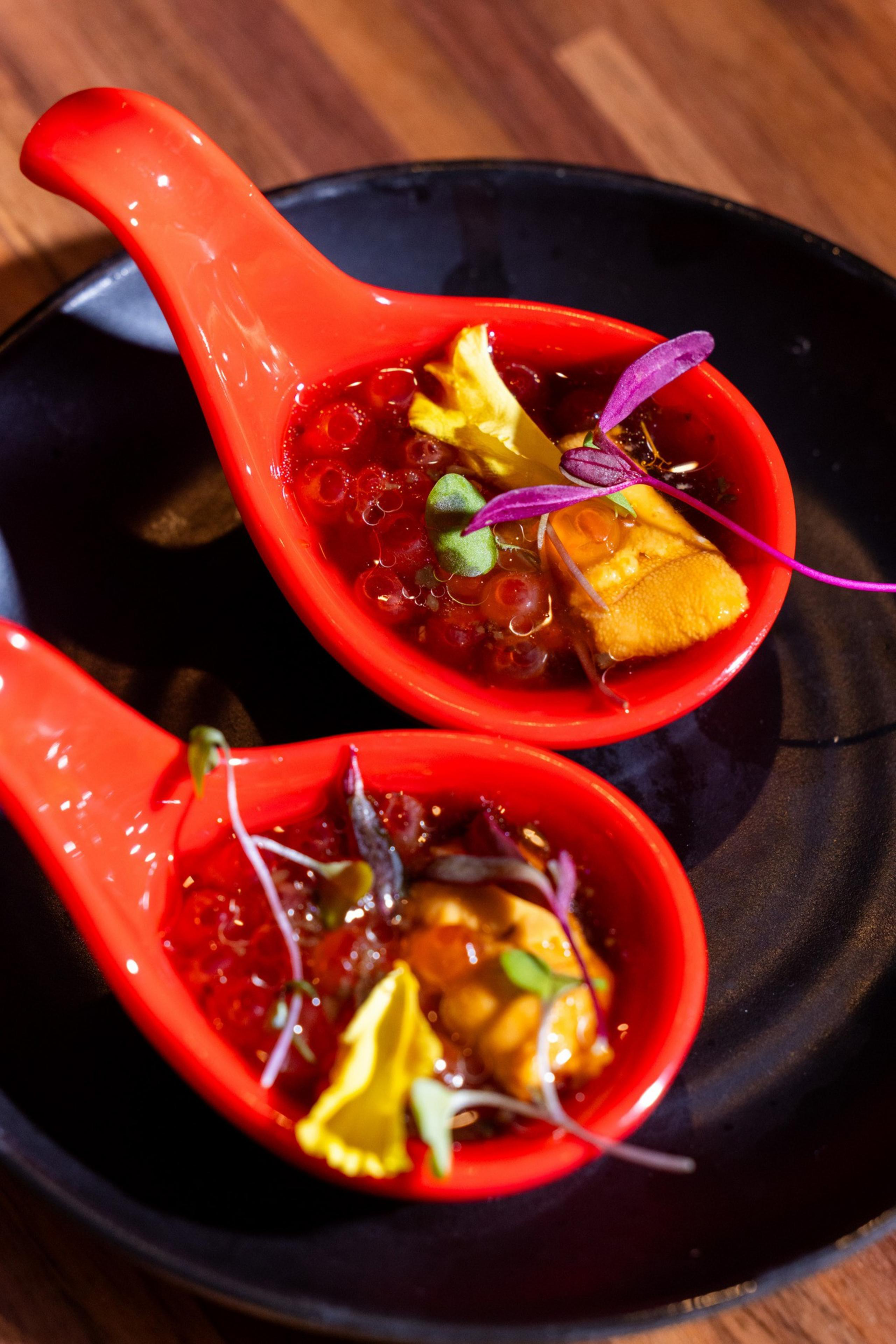 Two red spoons hold a vibrant dish with red spheres, garnished with yellow petals and microgreens, set on a dark plate on a wooden table.