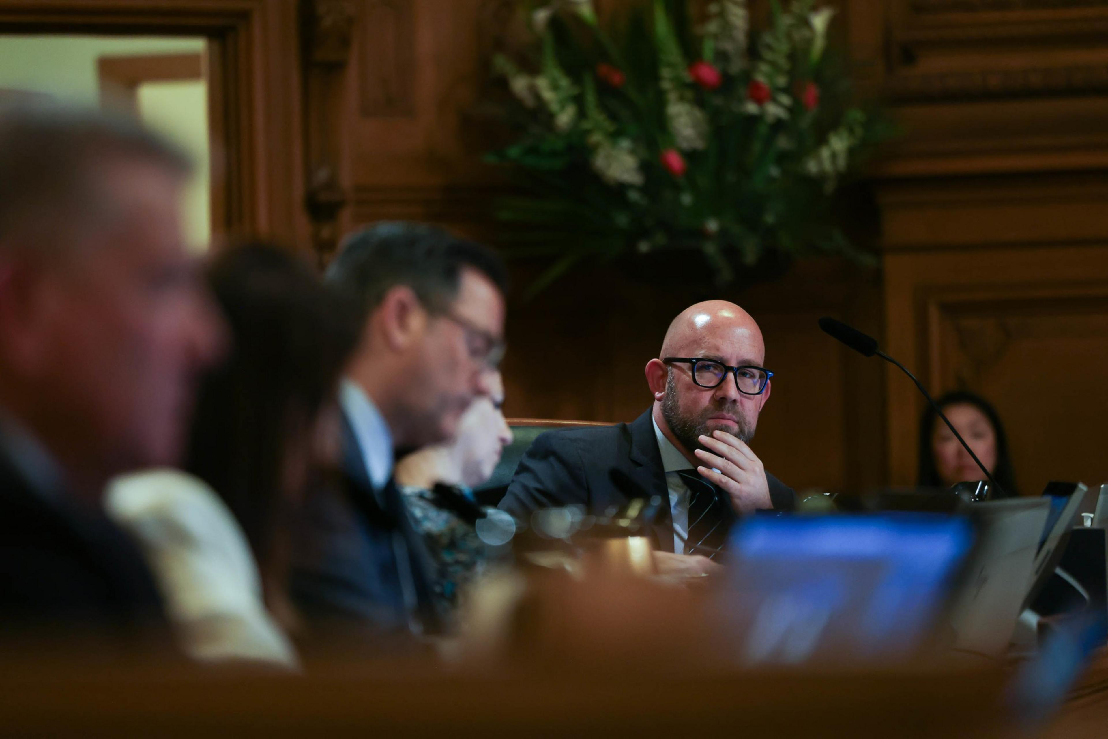 A man in glasses, focused and resting his chin on his hand, sits amidst blurred figures in a formal meeting room setting.