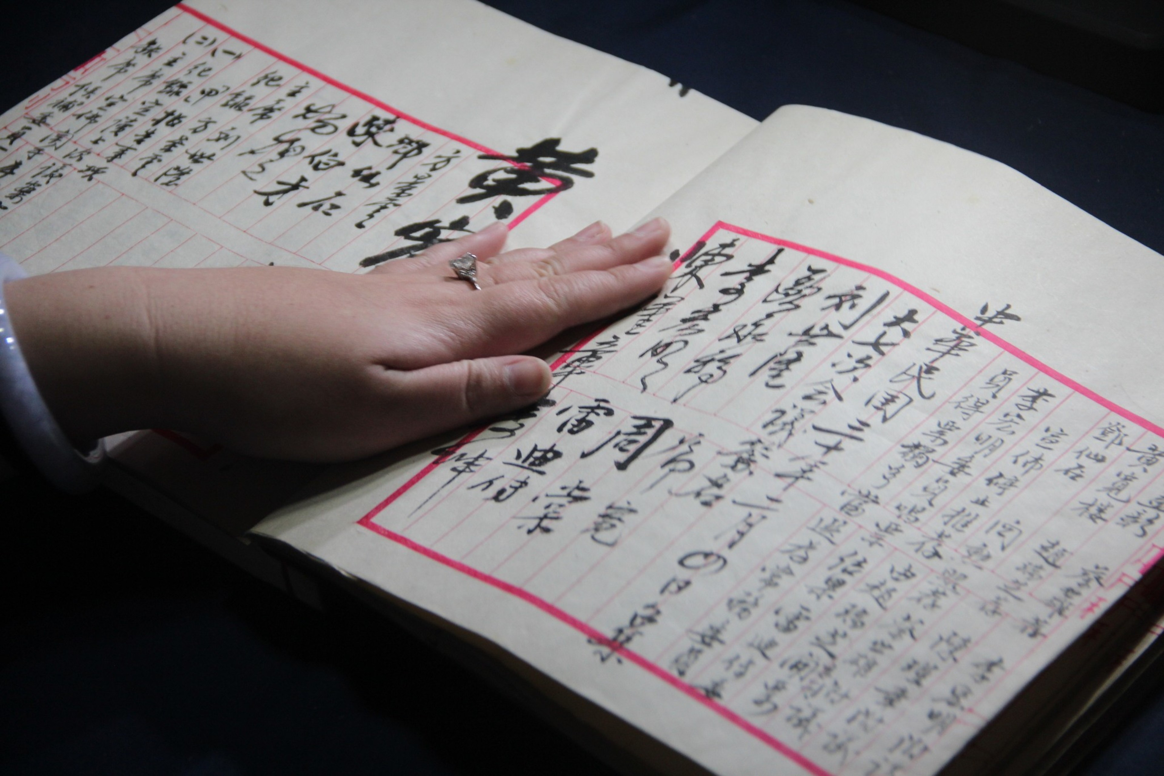 A hand rests on an open book with handwritten Chinese characters. The page margins are red, and the hand wears a ring and bracelet.