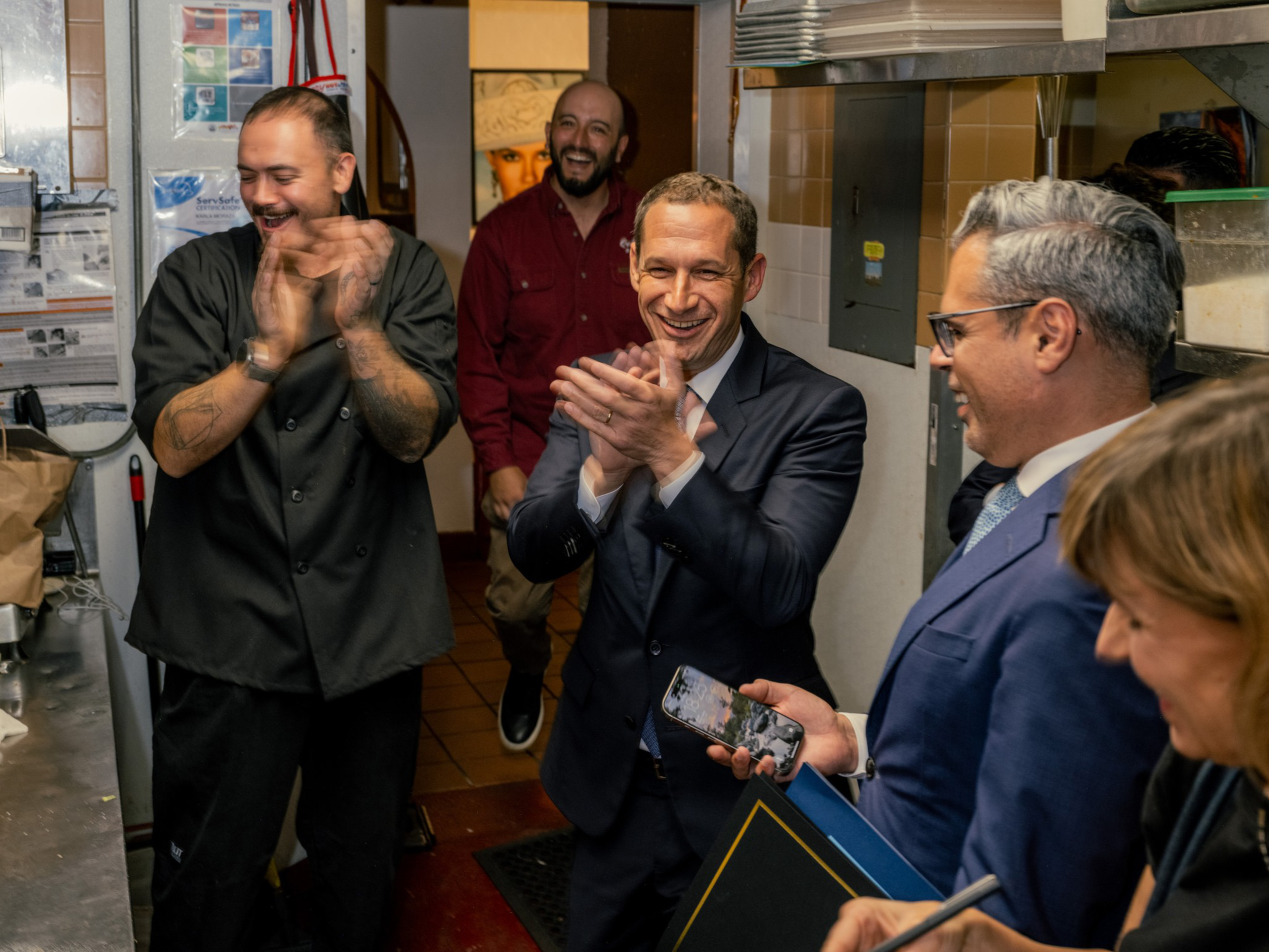 Four men and one woman smile and clap inside a kitchen, with one man holding a phone and another dressed in a chef’s coat.