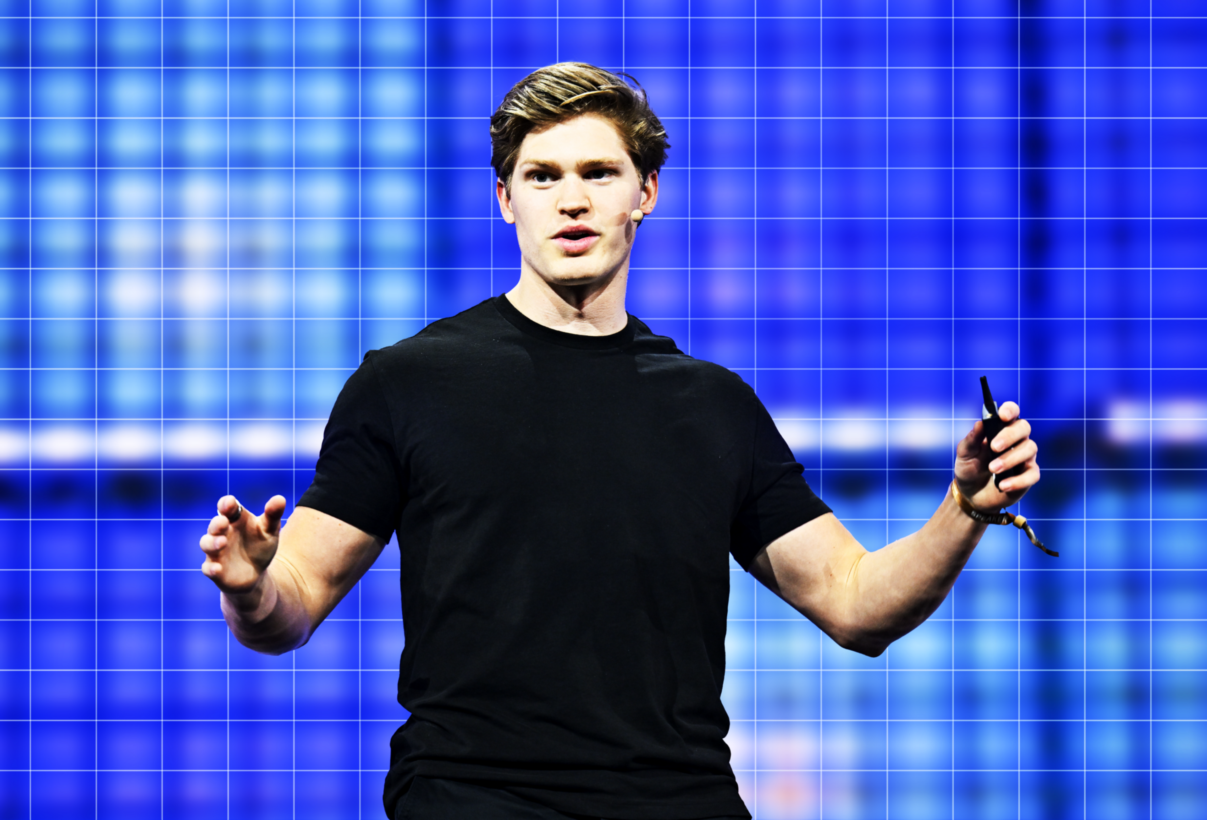 A young man in a black t-shirt speaks animatedly on stage, holding a small device with a blue grid-like background behind him.