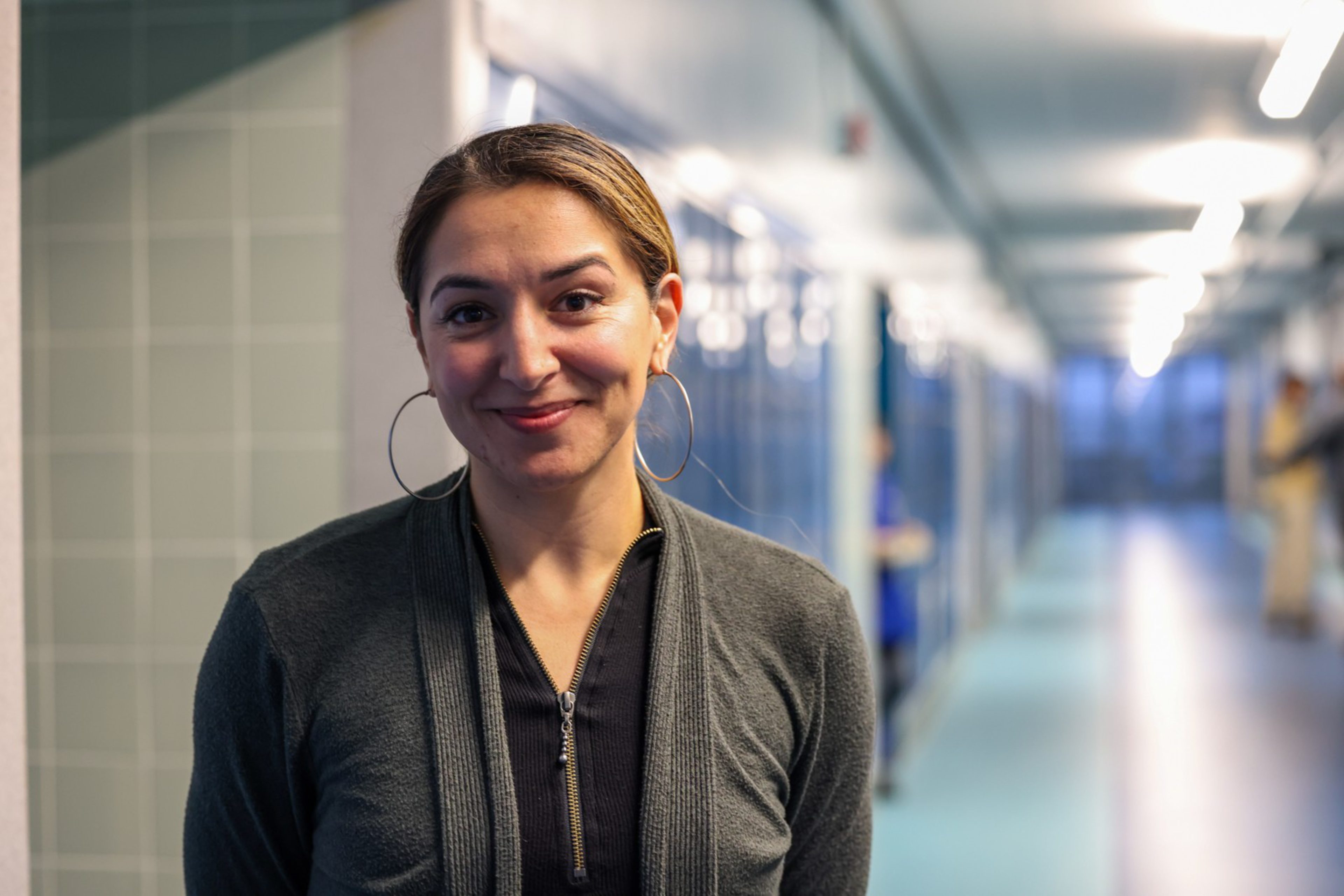 A woman with brown hair tied back, wearing large hoop earrings and a dark zip-up sweater, smiles gently in a brightly lit corridor.