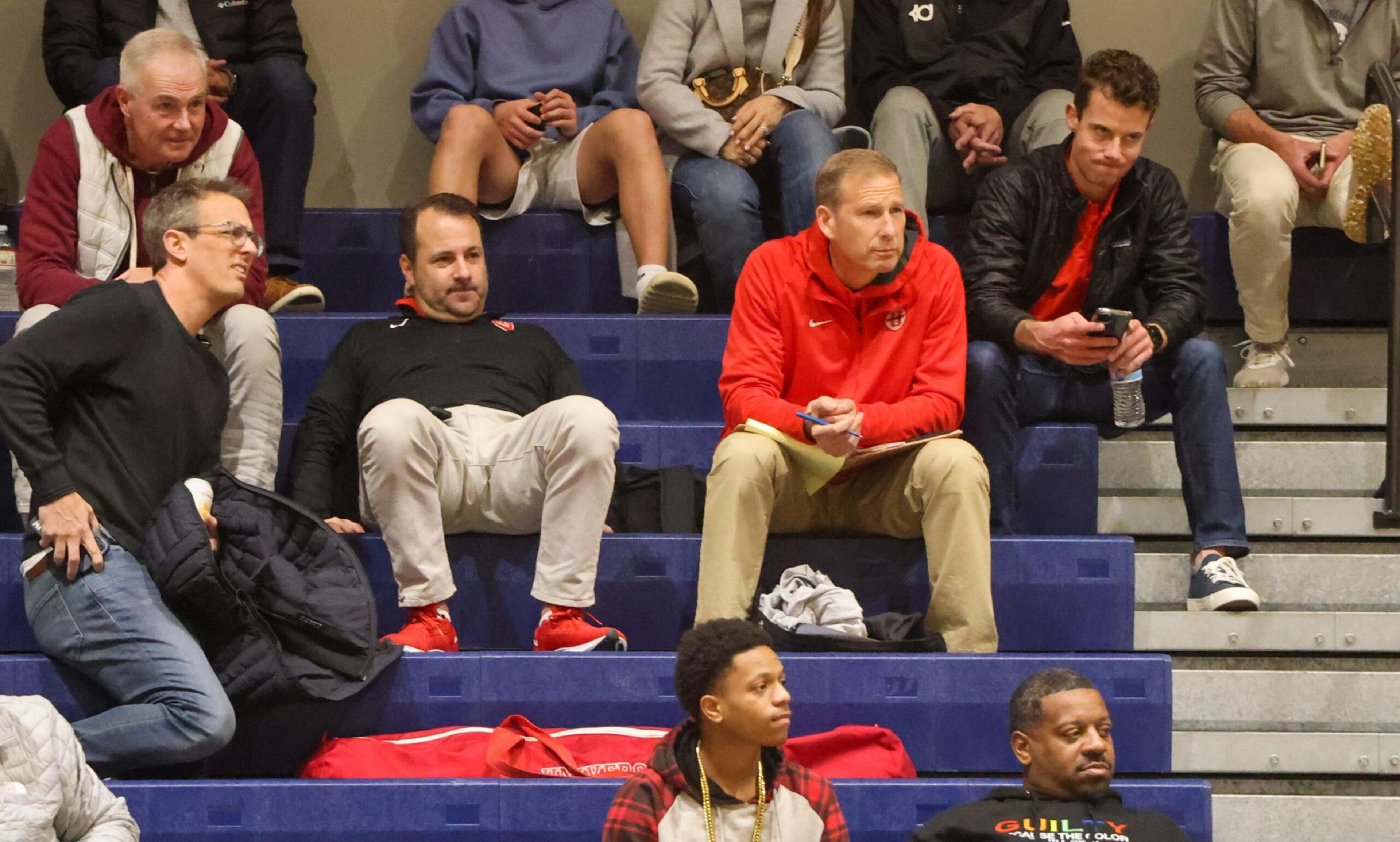 A group of men watch an event from blue bleachers, some sitting and some standing, with one holding a phone and another writing on a notepad.
