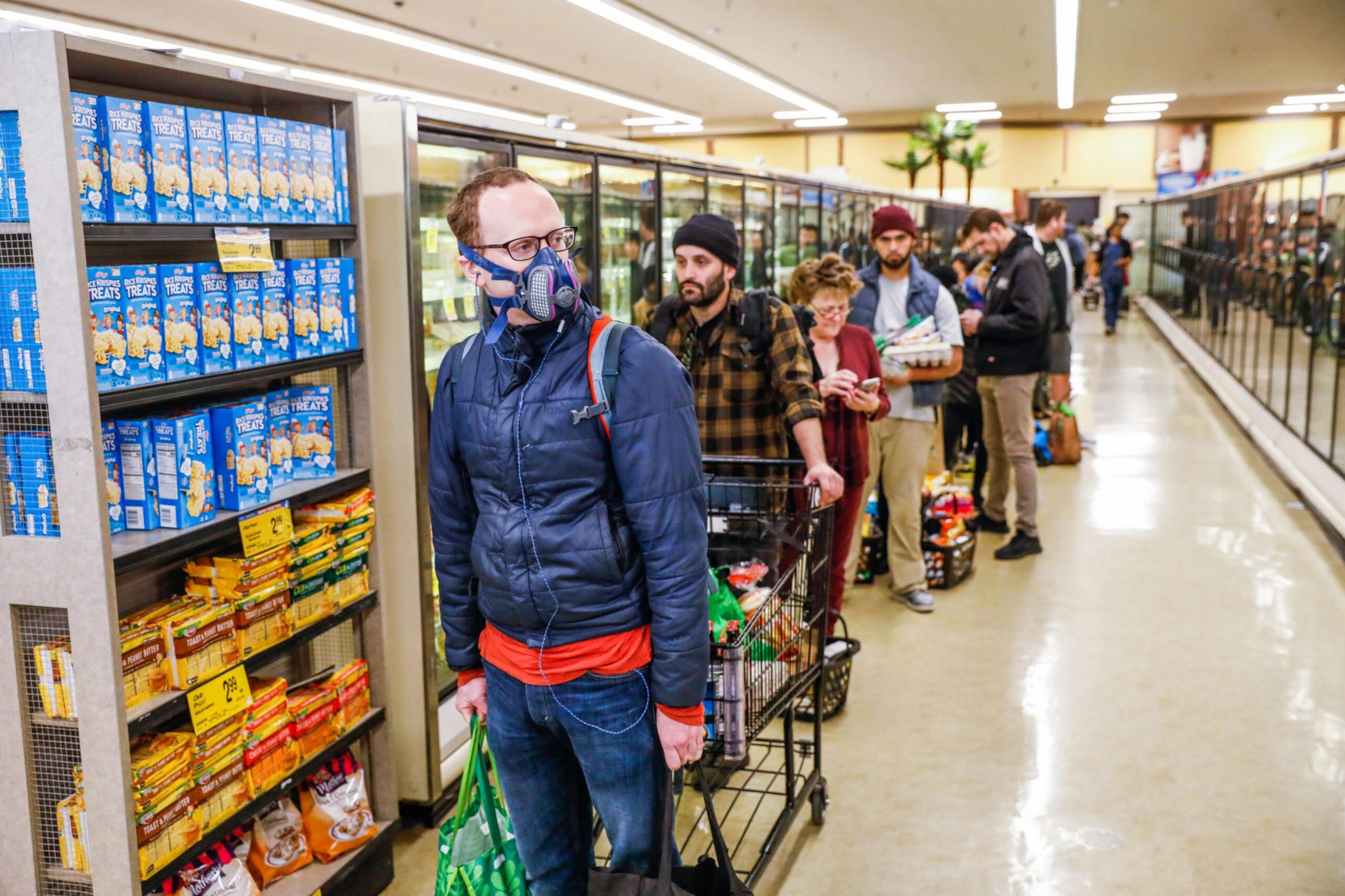 Several people stand in a long grocery store checkout line, with the person in front wearing a protective face mask and holding reusable bags.