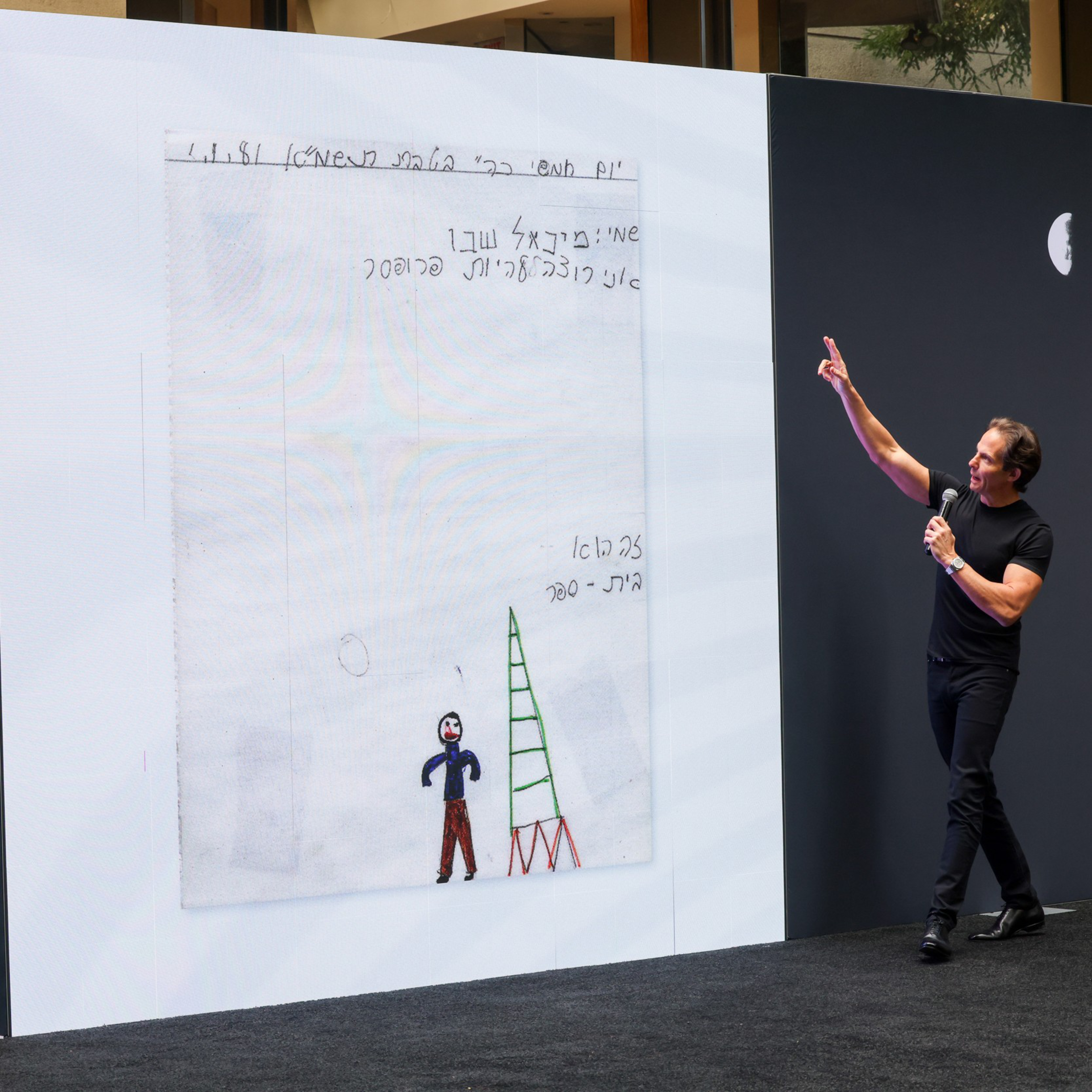 A man in black clothes holds a microphone and points at a large screen displaying a child-like drawing of a person and a green ladder.