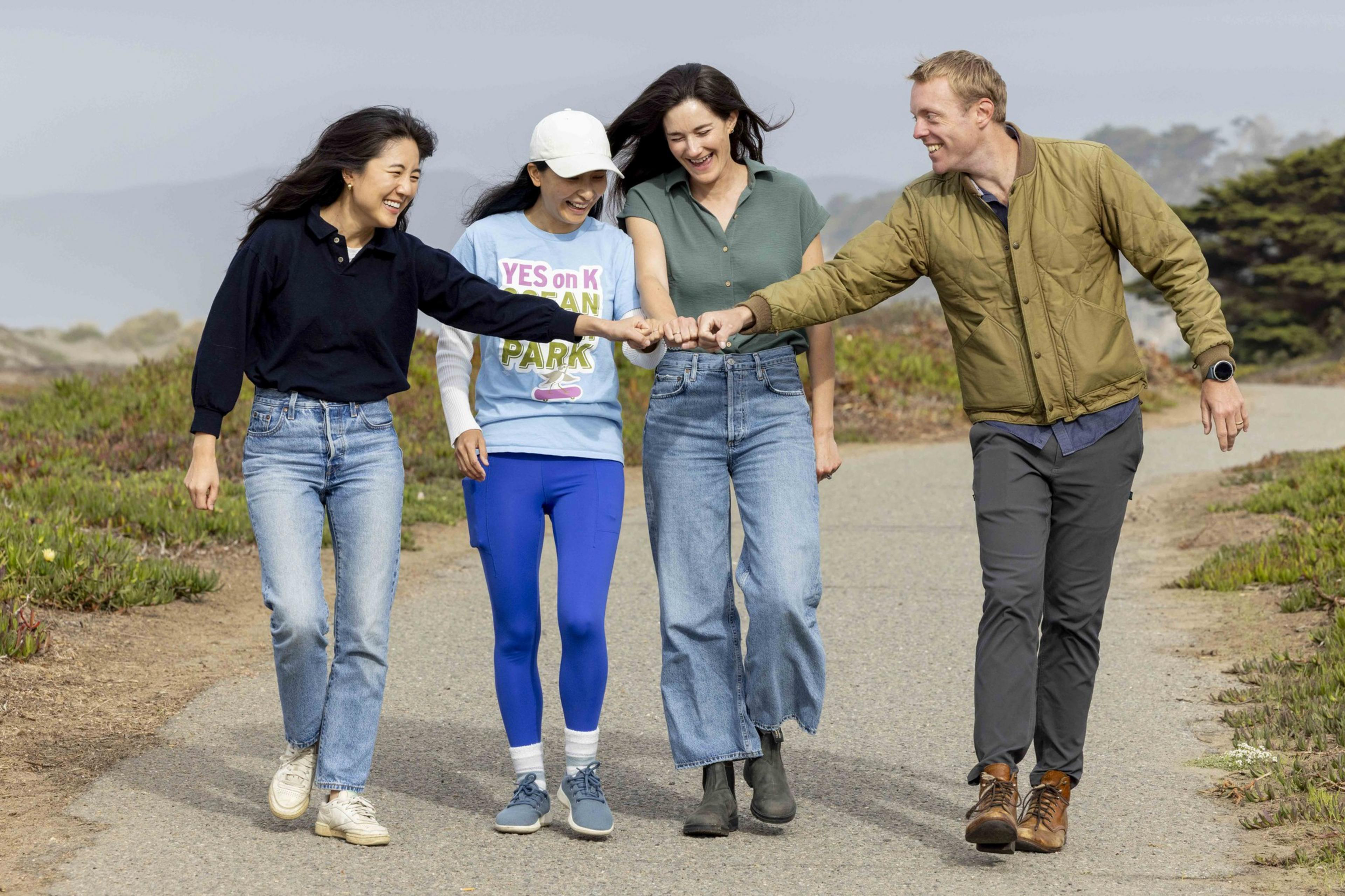Four people are walking and laughing on a path with greenery around. They're casually dressed, fist-bumping, and appear to be enjoying the moment.