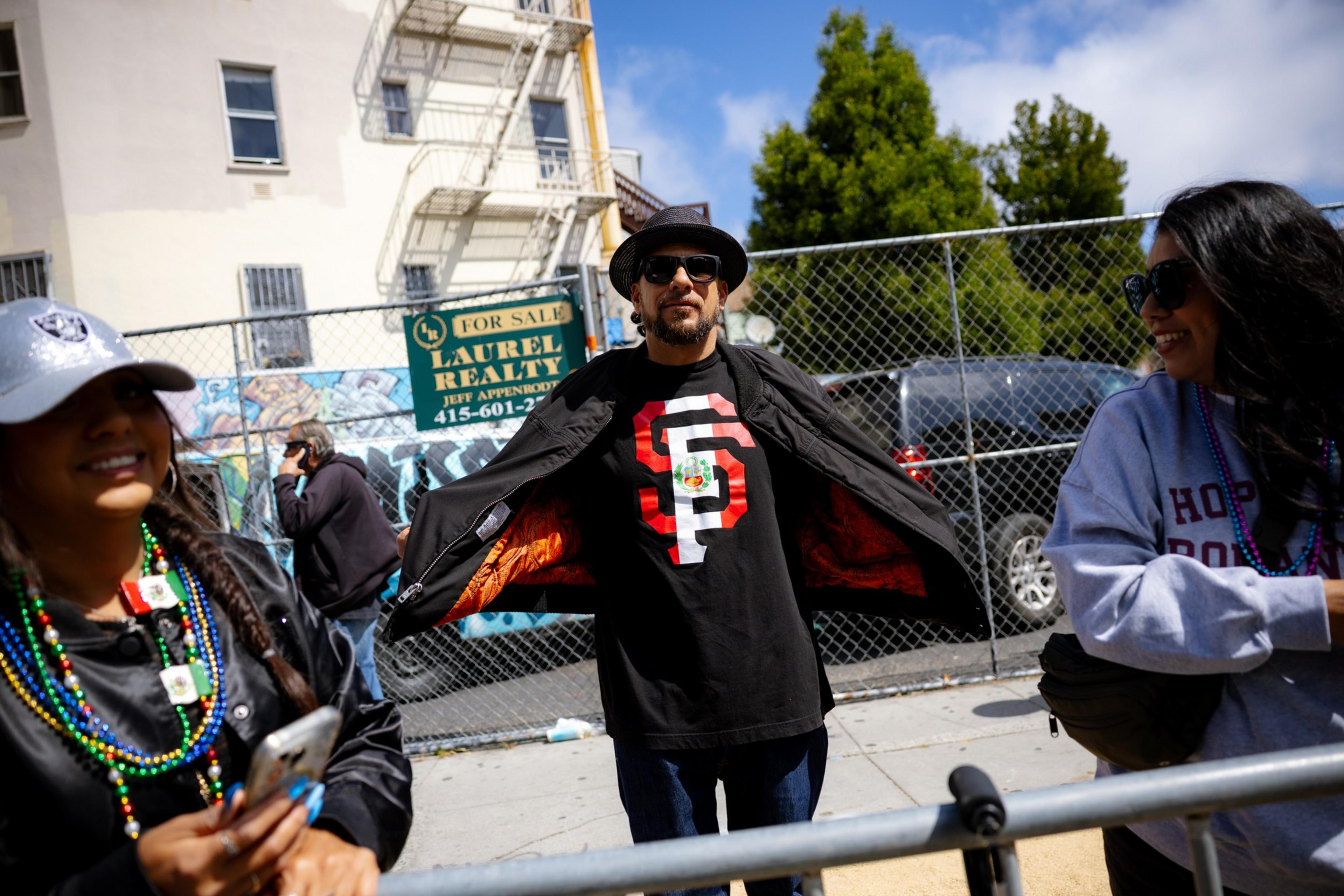 A person in a hat and sunglasses opens his jacket to reveal a shirt with large letters. Two smiling people, one holding a phone, stand nearby on a sunny day.