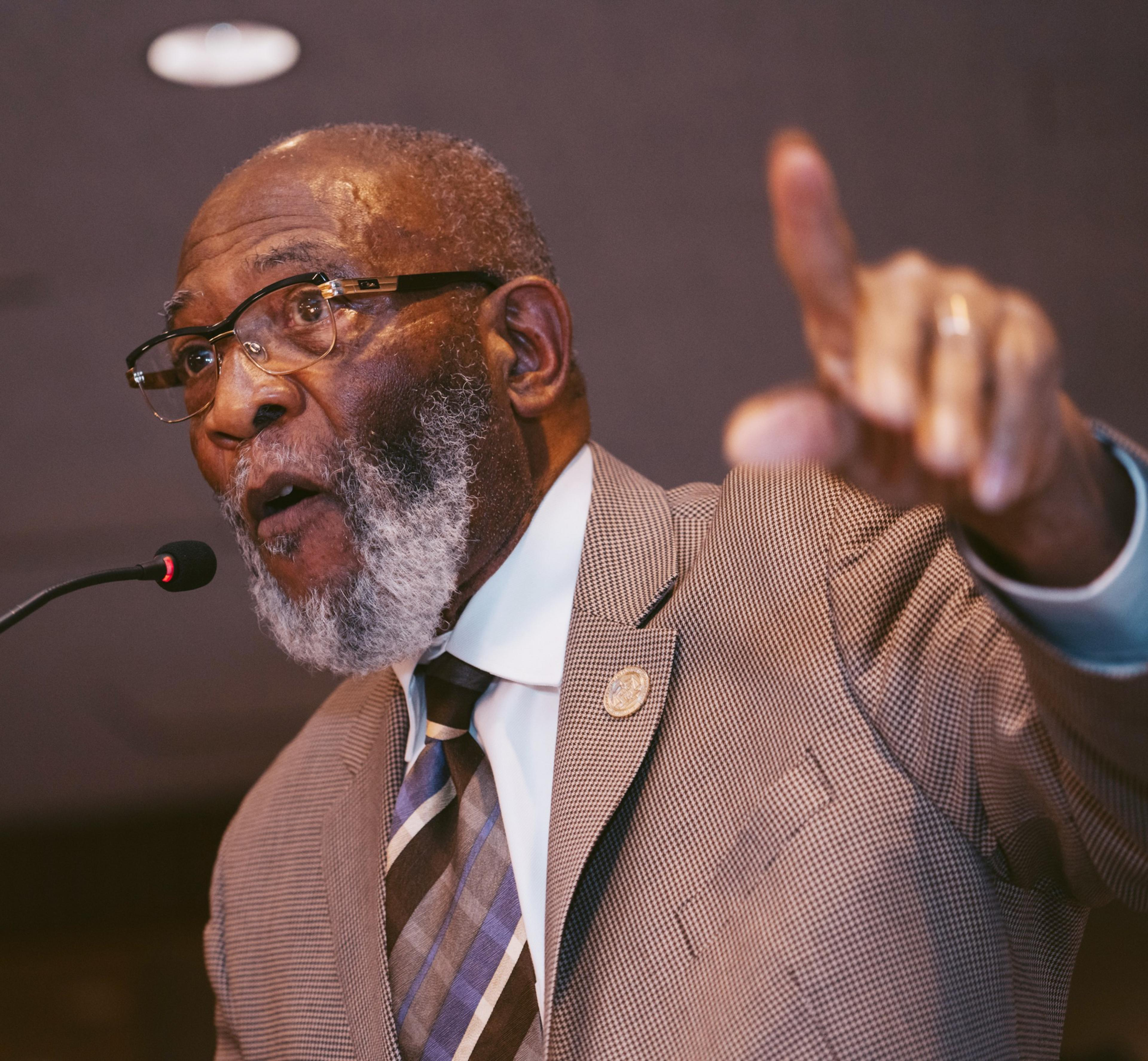 An elder man is speaking at a microphone, pointing forward, wearing glasses, a suit, and a tie. He appears engaged in his topic.
