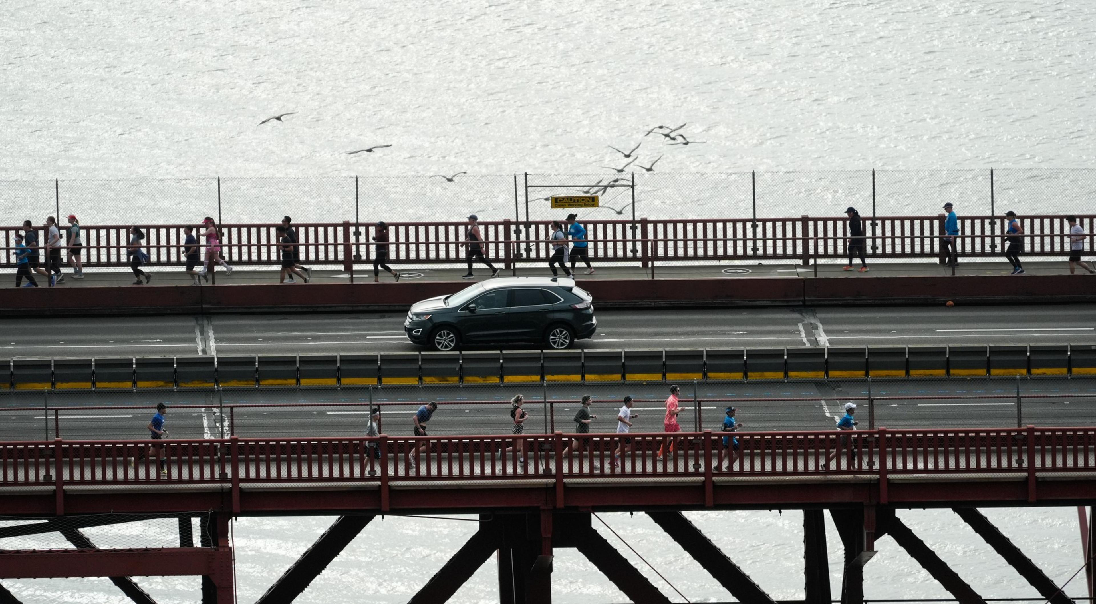 People are walking on two levels of a red bridge, separated by a road with a black SUV. Seagulls are flying in the background.