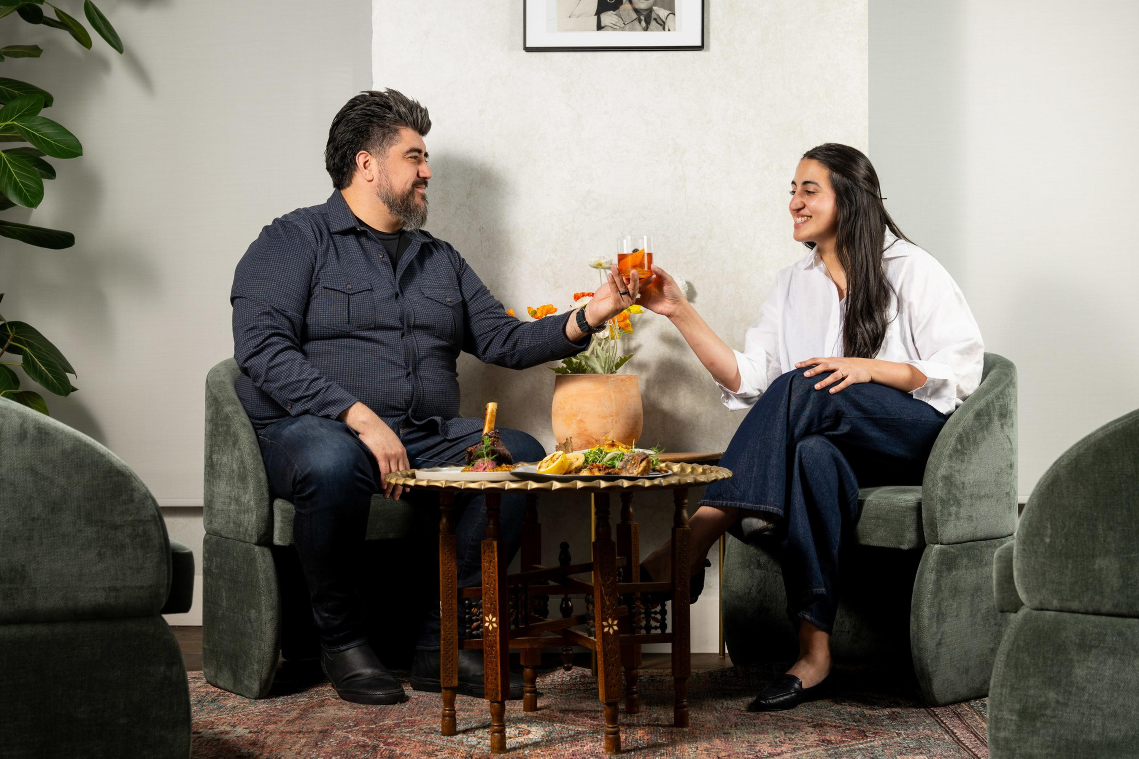A man and woman sit in green chairs, toasting with drinks across a small table adorned with food and flowers.