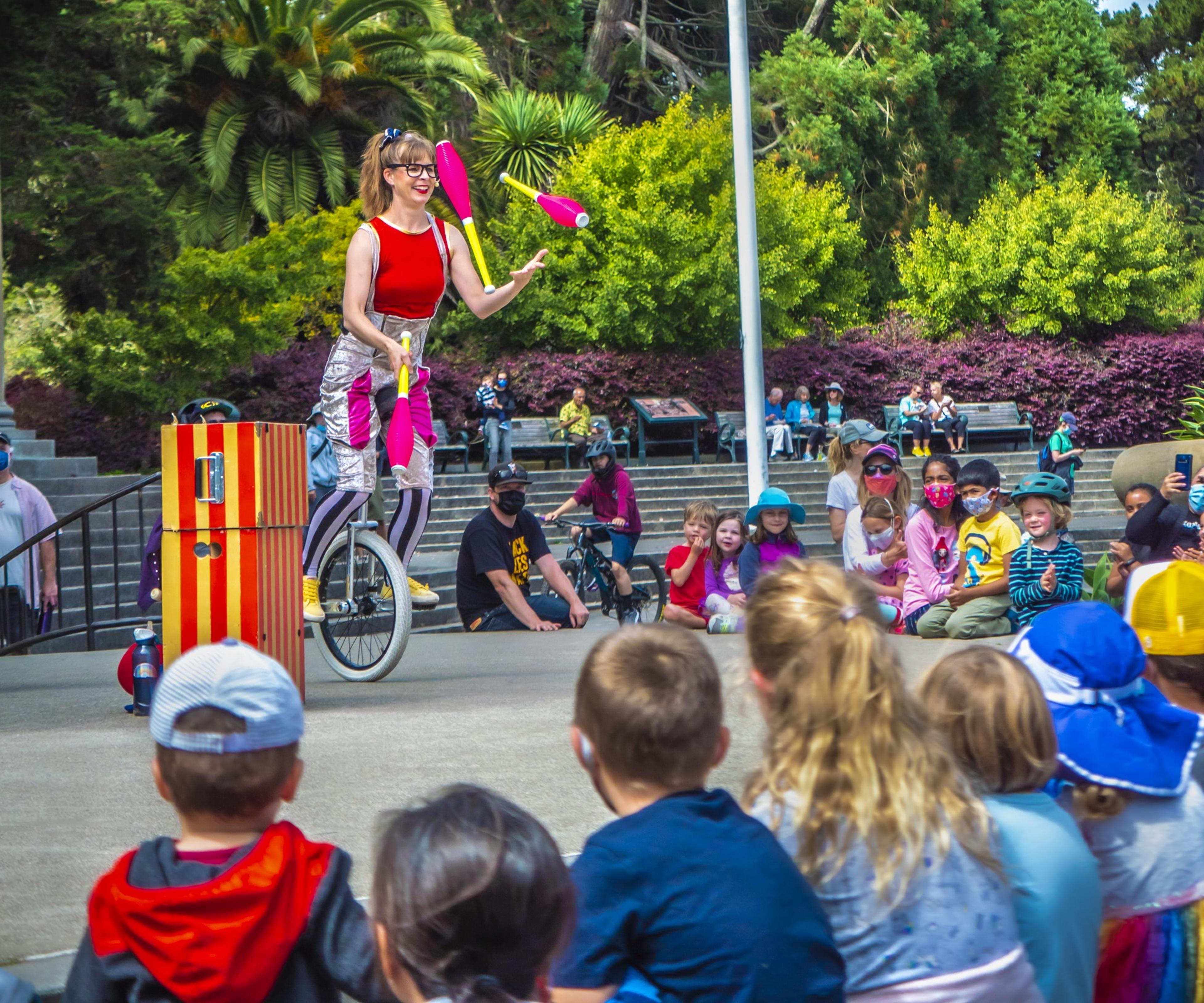 A performer juggles clubs while riding a unicycle before an audience of children and adults in a park, with lush greenery in the background.