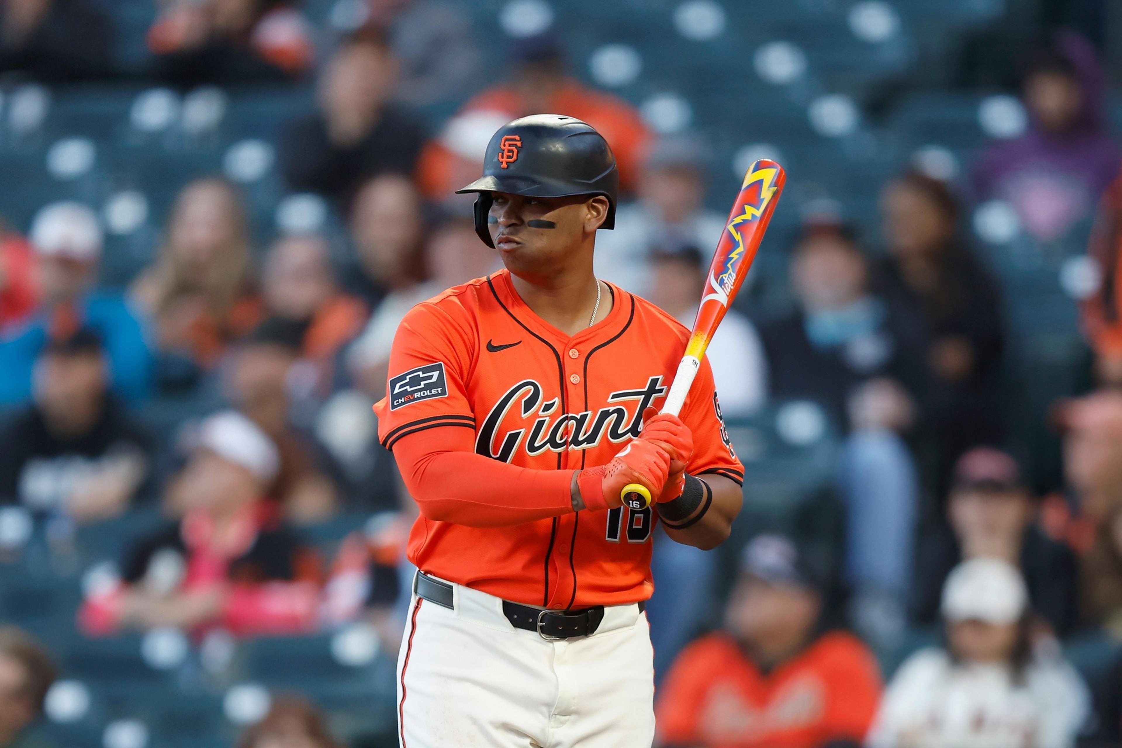A baseball player in a San Francisco Giants uniform grips a colorful bat, standing focused on the field with a crowd blurred in the background.