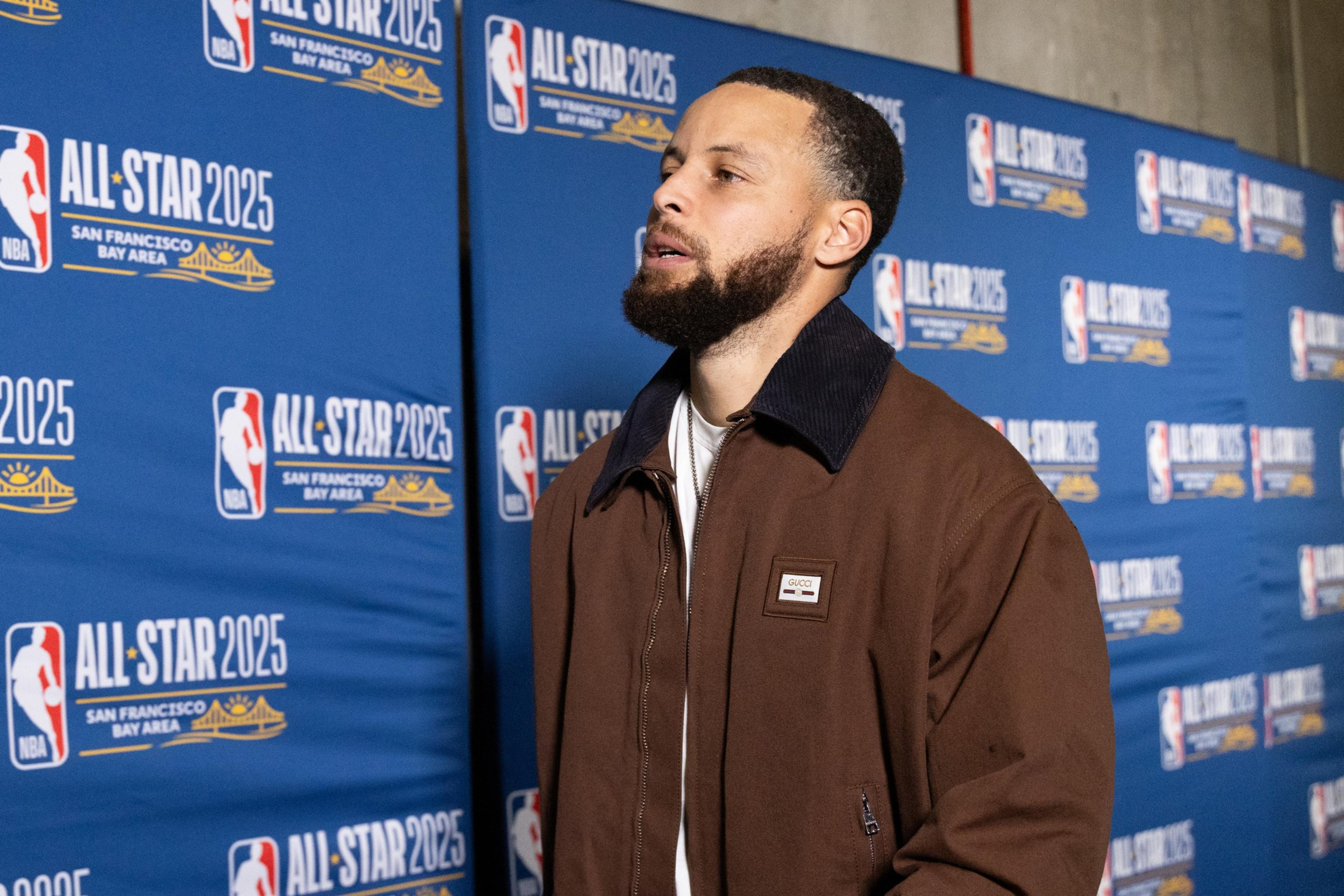 A person in a brown jacket stands in front of a blue "All-Star 2025 San Francisco Bay Area" backdrop with NBA logos.