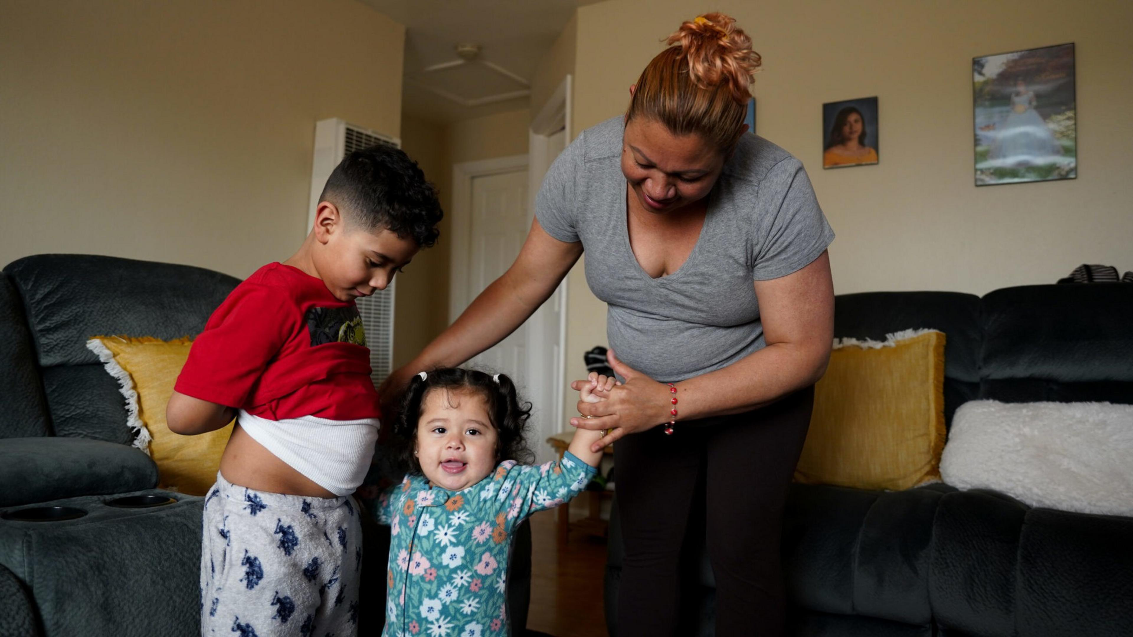 A woman helps a toddler in floral pajamas stand while a boy in a red shirt and pajama pants looks on in a living room with dark couches and yellow pillows.
