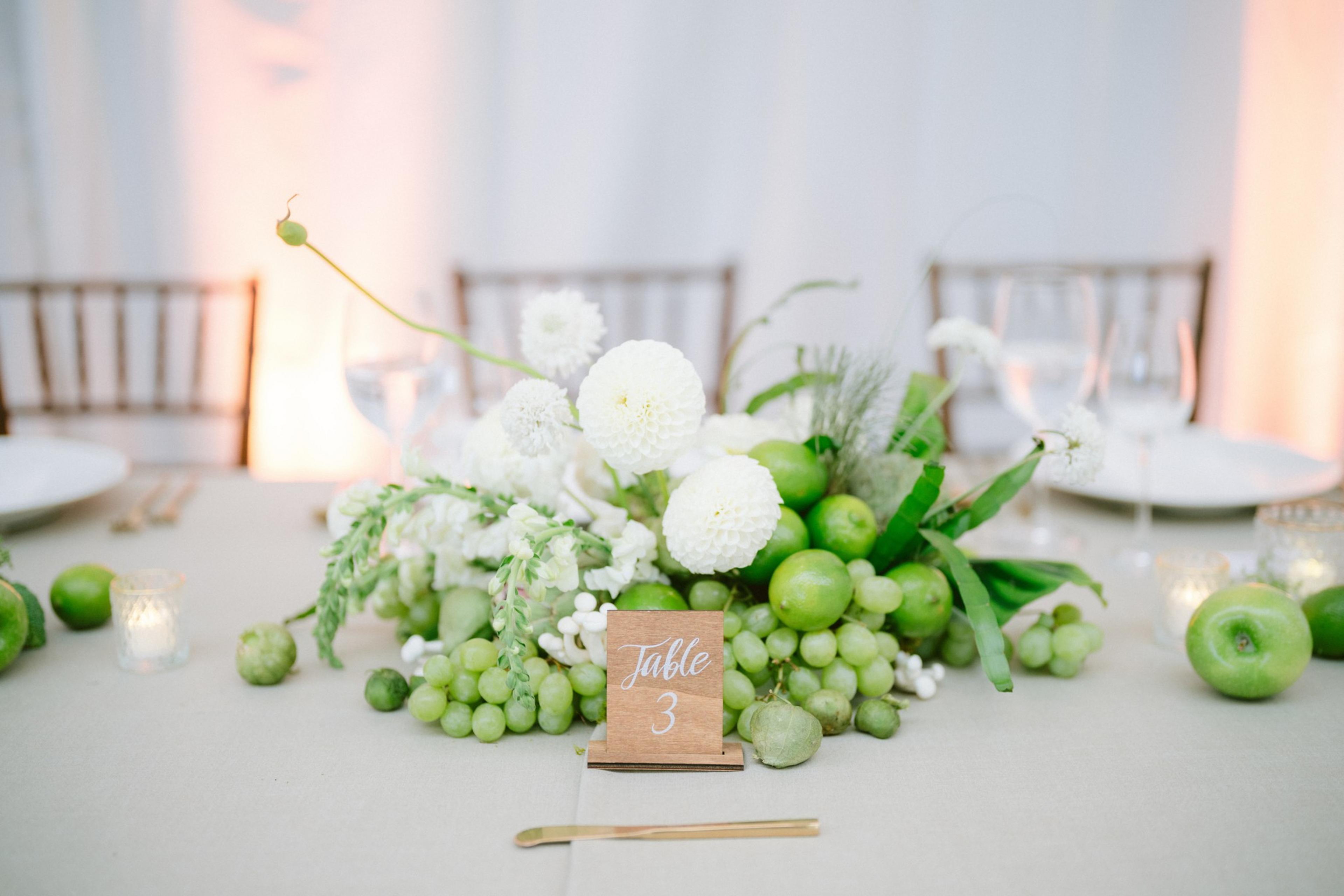 A centerpiece with white flowers, green grapes, and green apples sits on a table with a “Table 3" wooden sign, gold cutlery, and wine glasses nearby.