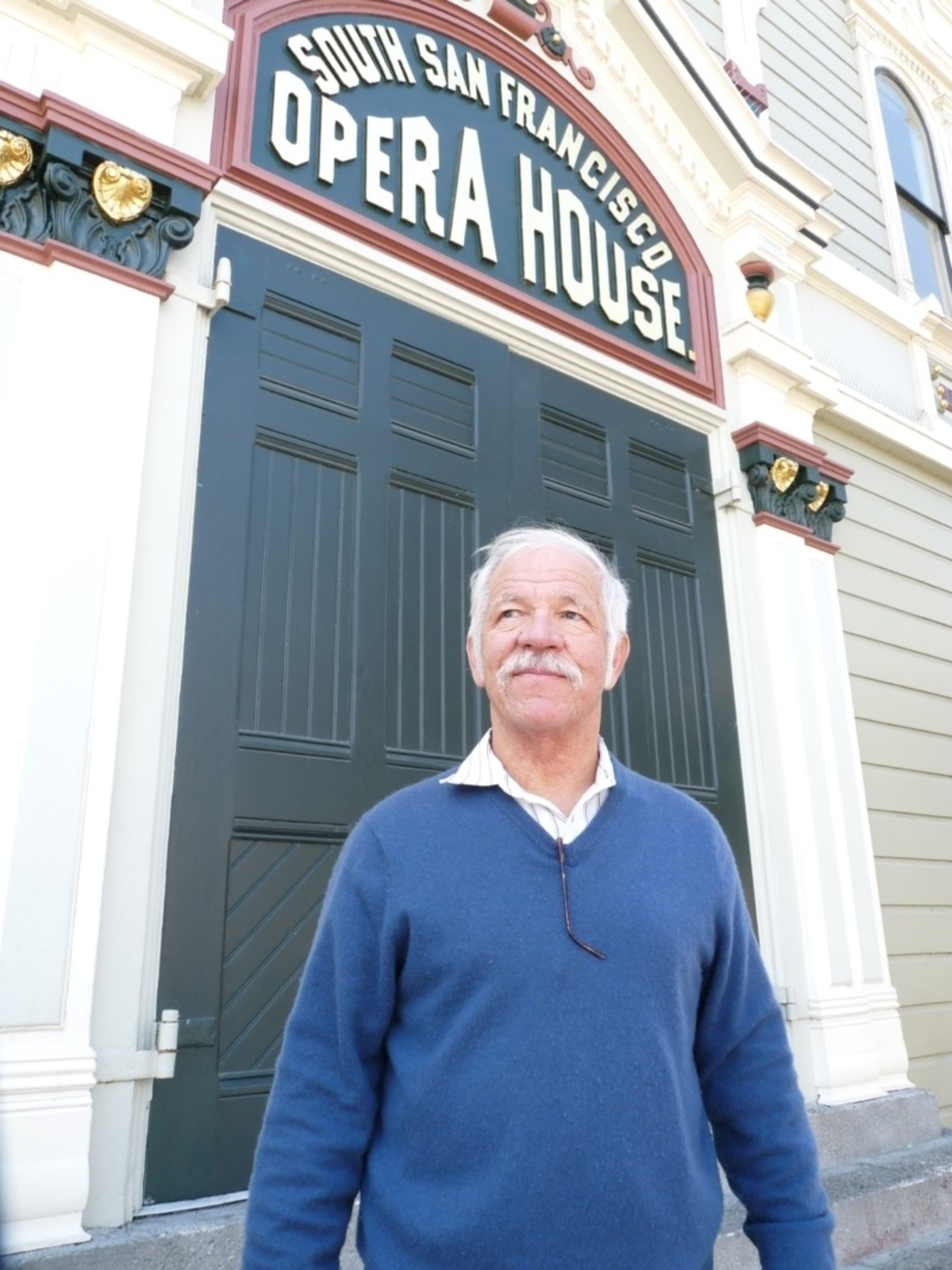 An older man with white hair and a mustache stands in a blue sweater outside the South San Francisco Opera House with large dark green doors behind him.