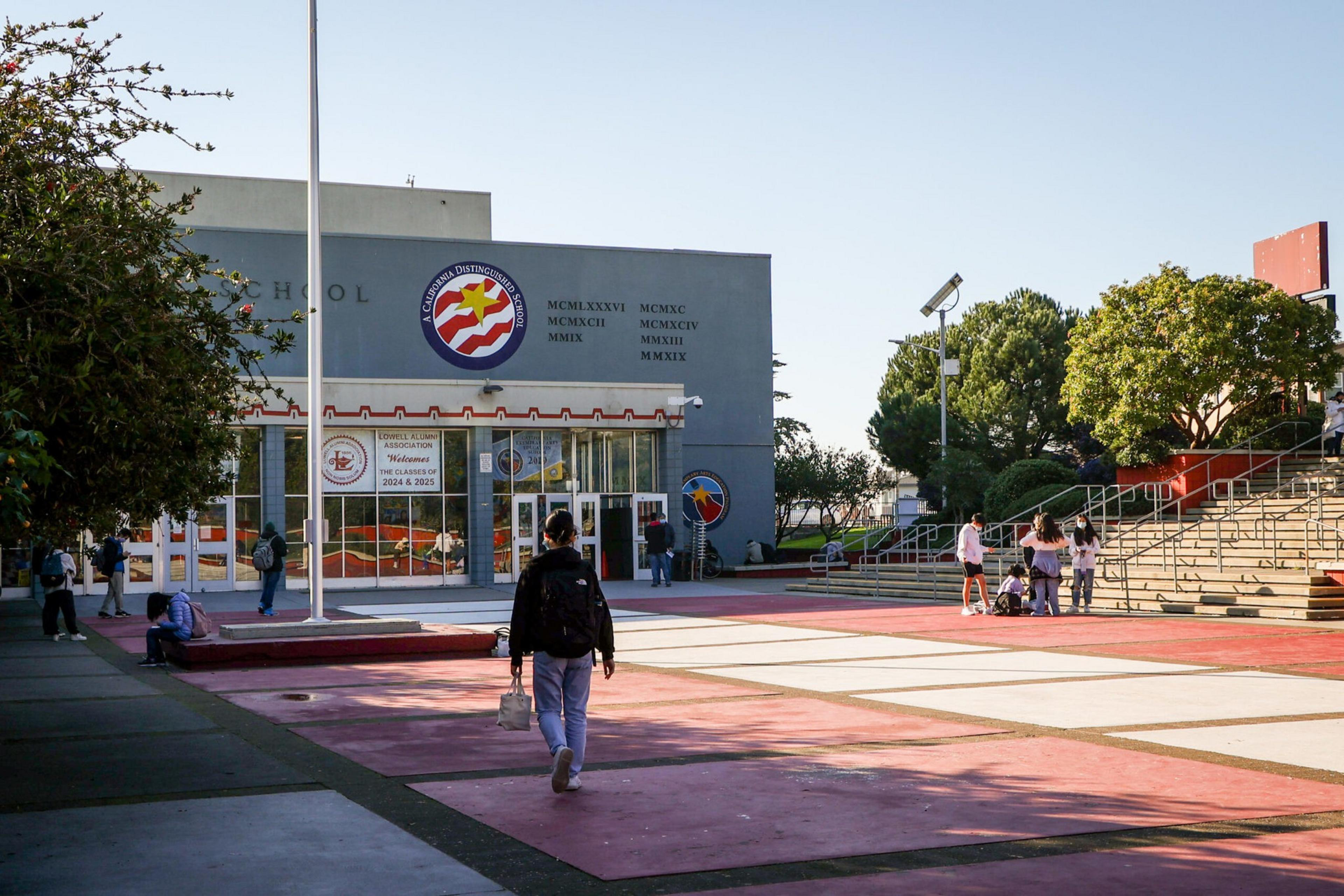 Students walk and gather outside a modern school building with large windows, surrounded by trees and outdoor seating areas.