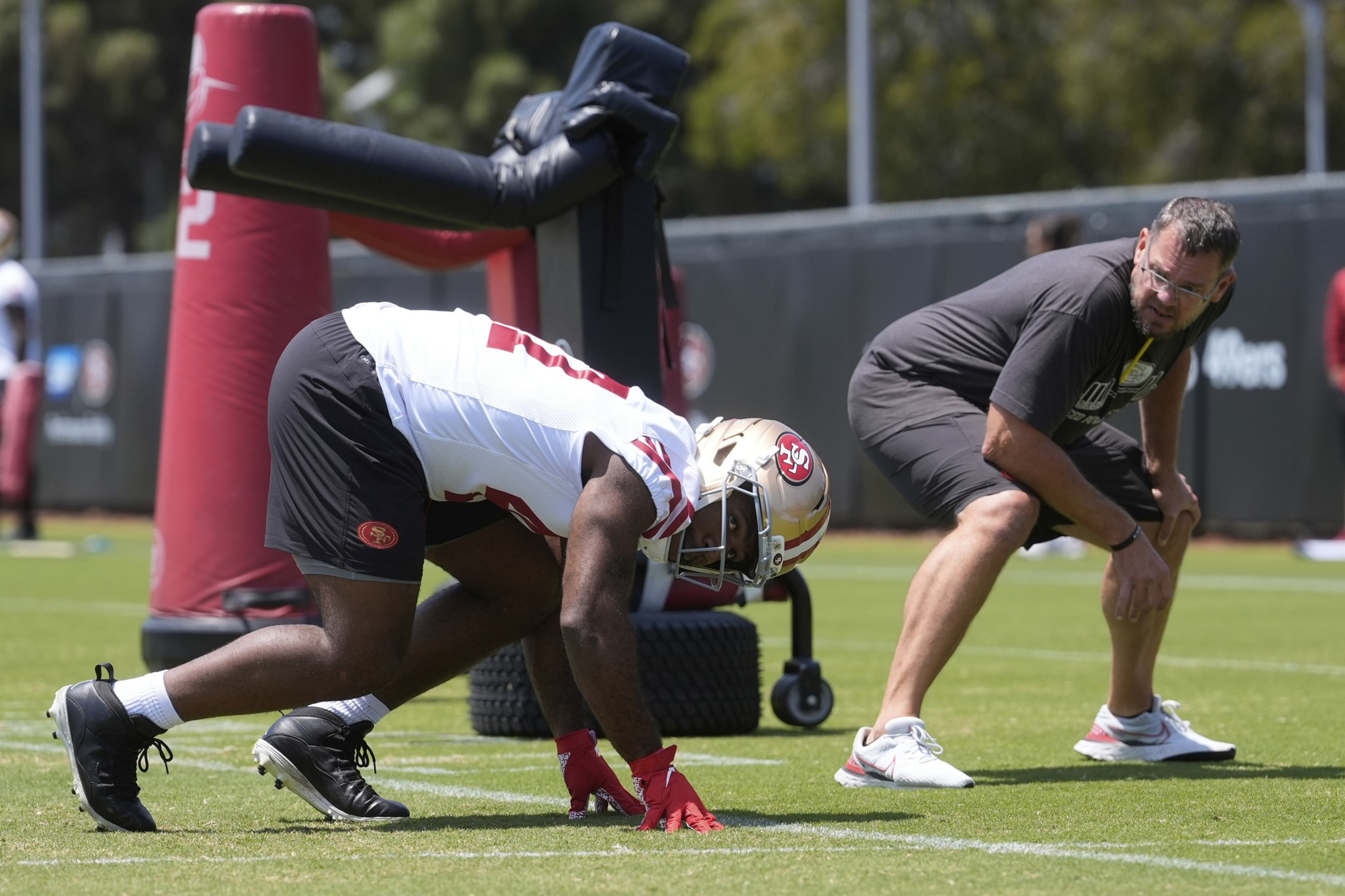 A football player in full gear, wearing a helmet, crouches in a ready stance on a grassy field. A coach observes nearby, both focused on practice.
