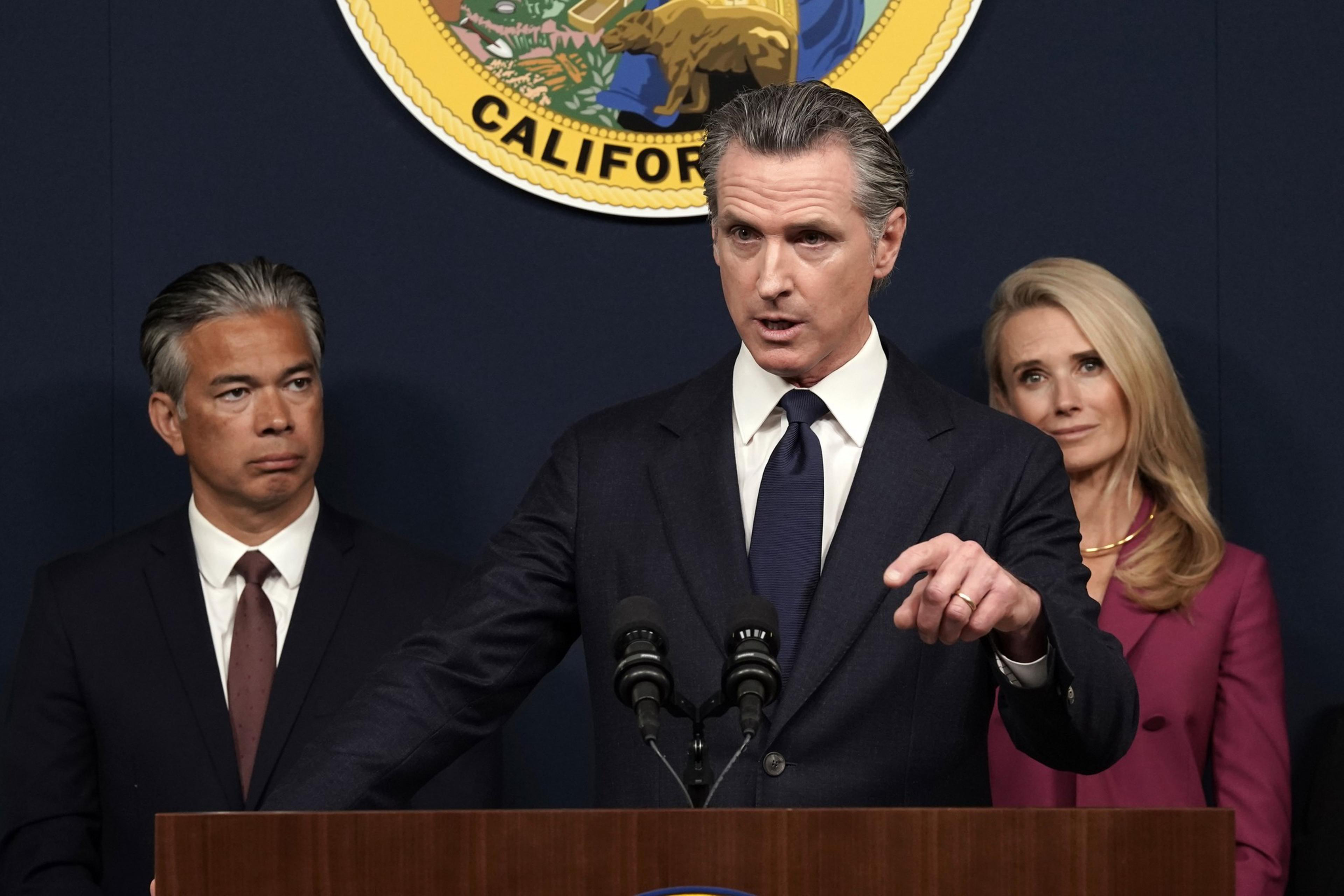Man and woman look on as another man speaks in front of a podium