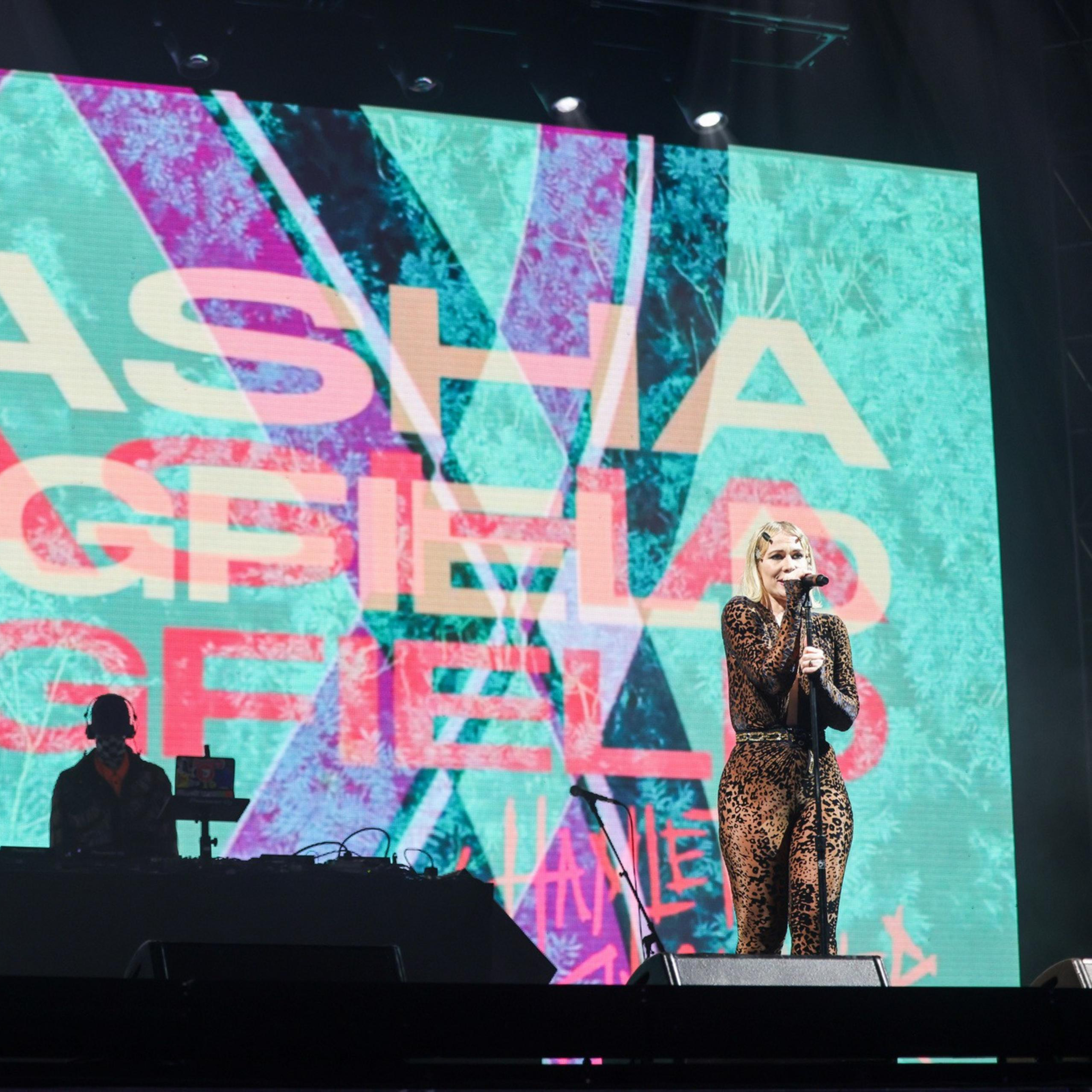 A performer in a leopard-print outfit sings into a microphone on stage. A large screen behind displays colorful graphics and the text &quot;NATASHA BEDINGFIELD.&quot;