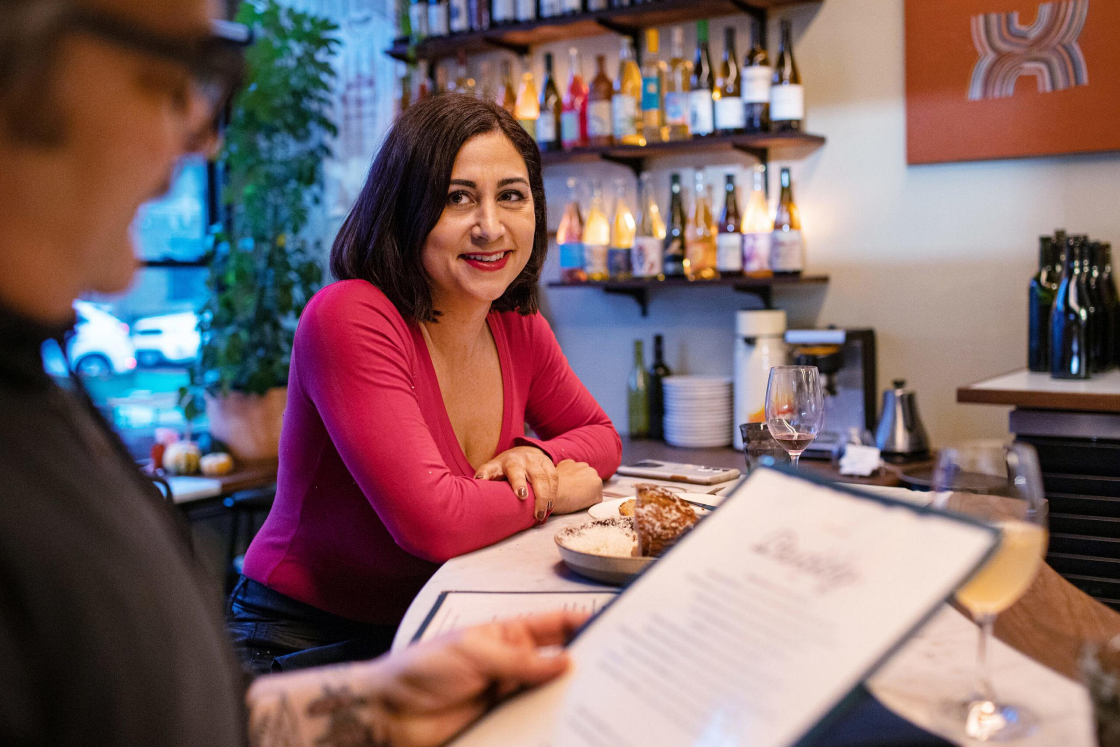 A woman in a red top smiles while sitting at a bar with food and drinks, as a man with glasses reads a menu in the foreground.