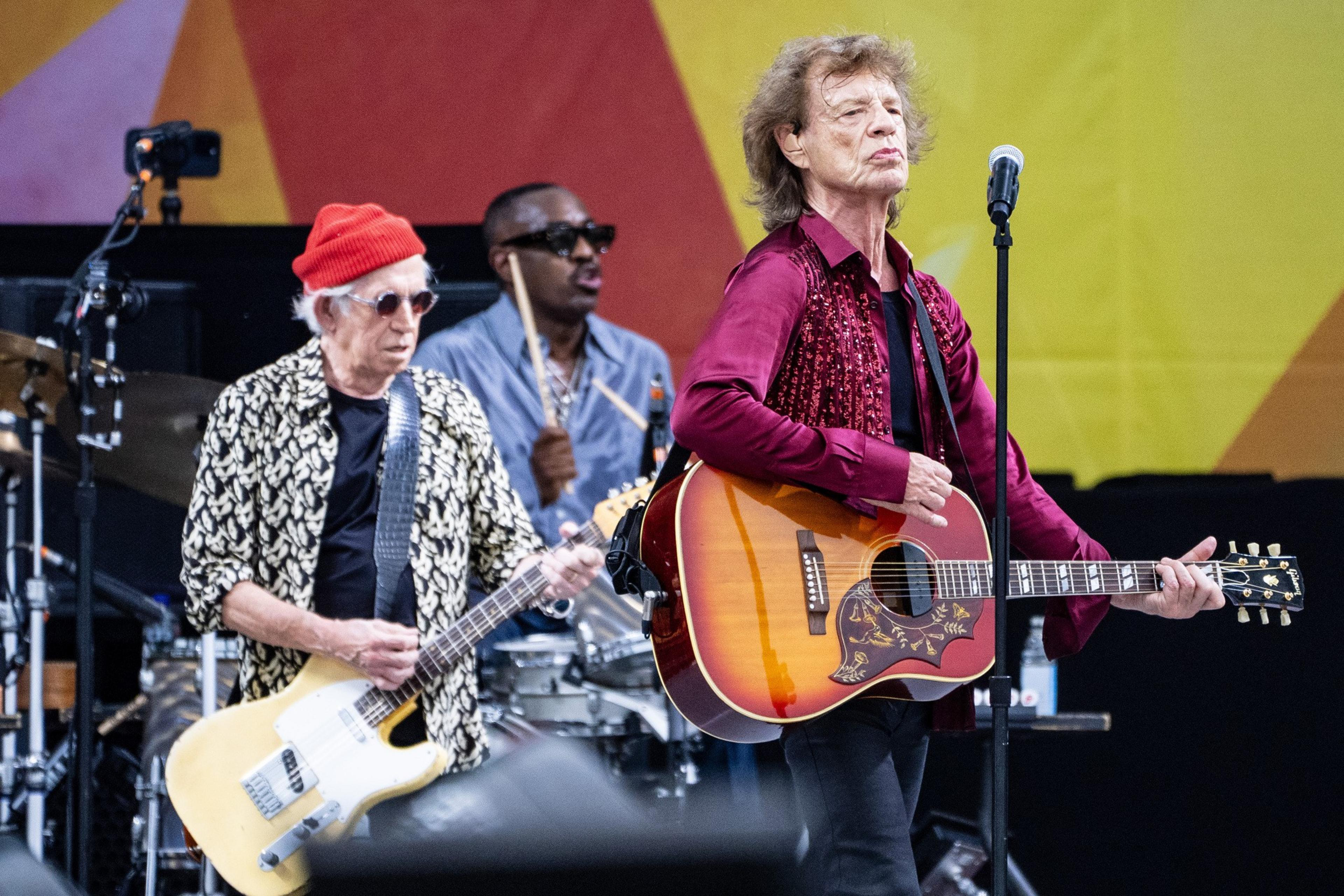 Three musicians perform on stage: one with a yellow electric guitar and red beanie, another with a tambourine, and the lead singer playing an acoustic guitar near a microphone.