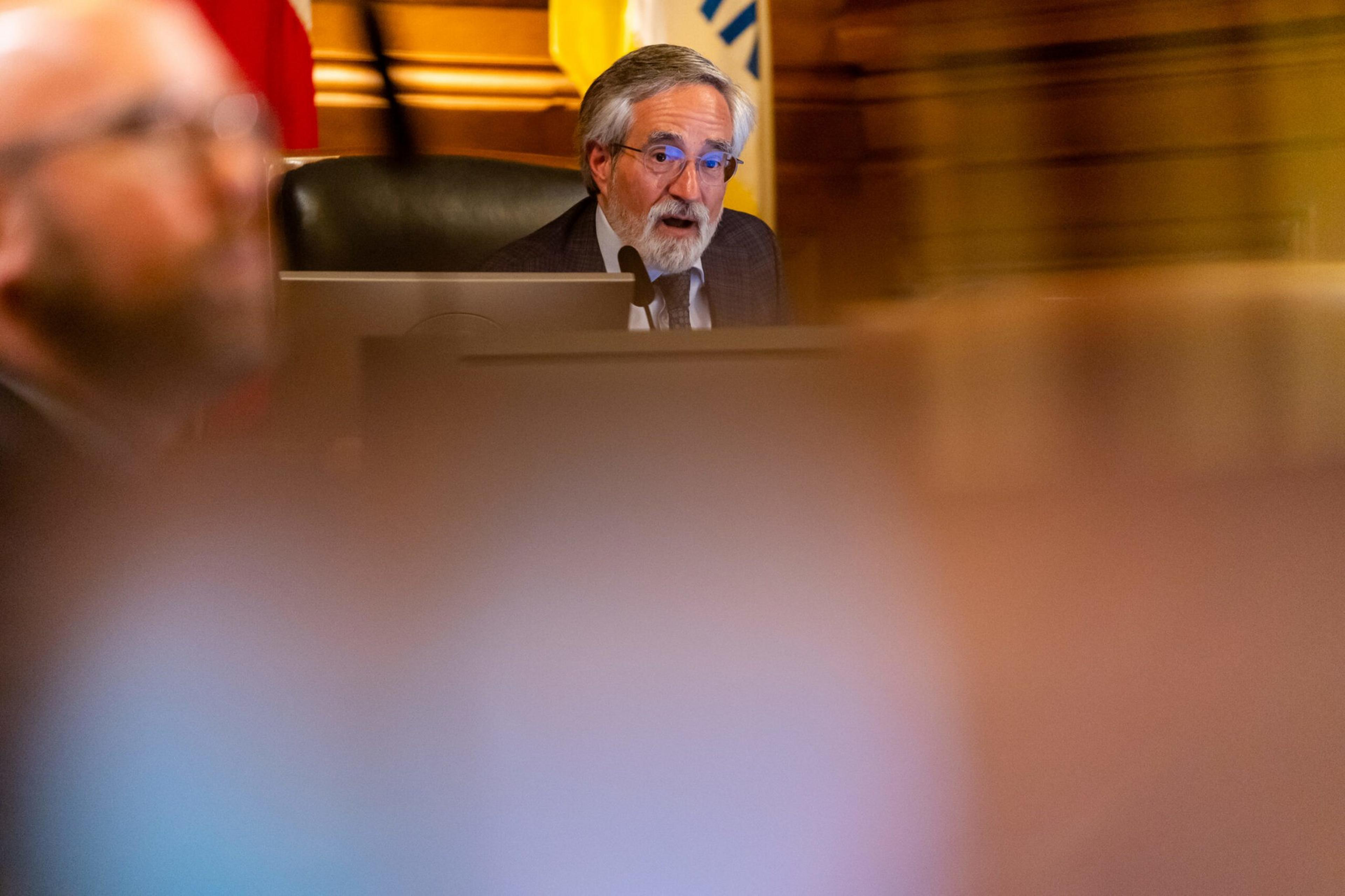 An older man with gray hair and glasses speaks behind a desk with a microphone, partially obscured by blurred foreground figures.