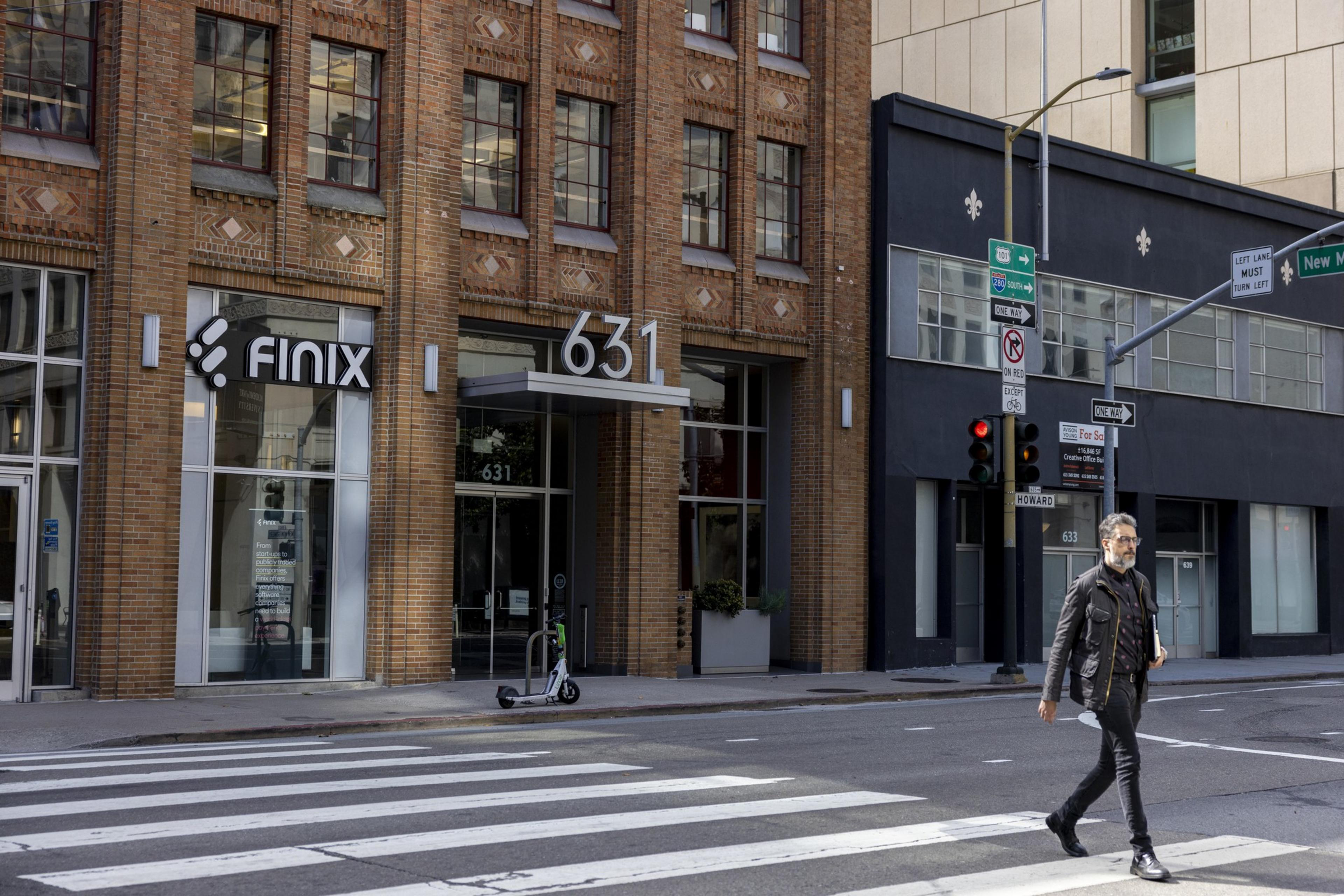 A man in a black jacket crosses a city street at a crosswalk. Behind him is a brick building with &quot;FINIX&quot; signage and the number &quot;631&quot; displayed above the entrance.