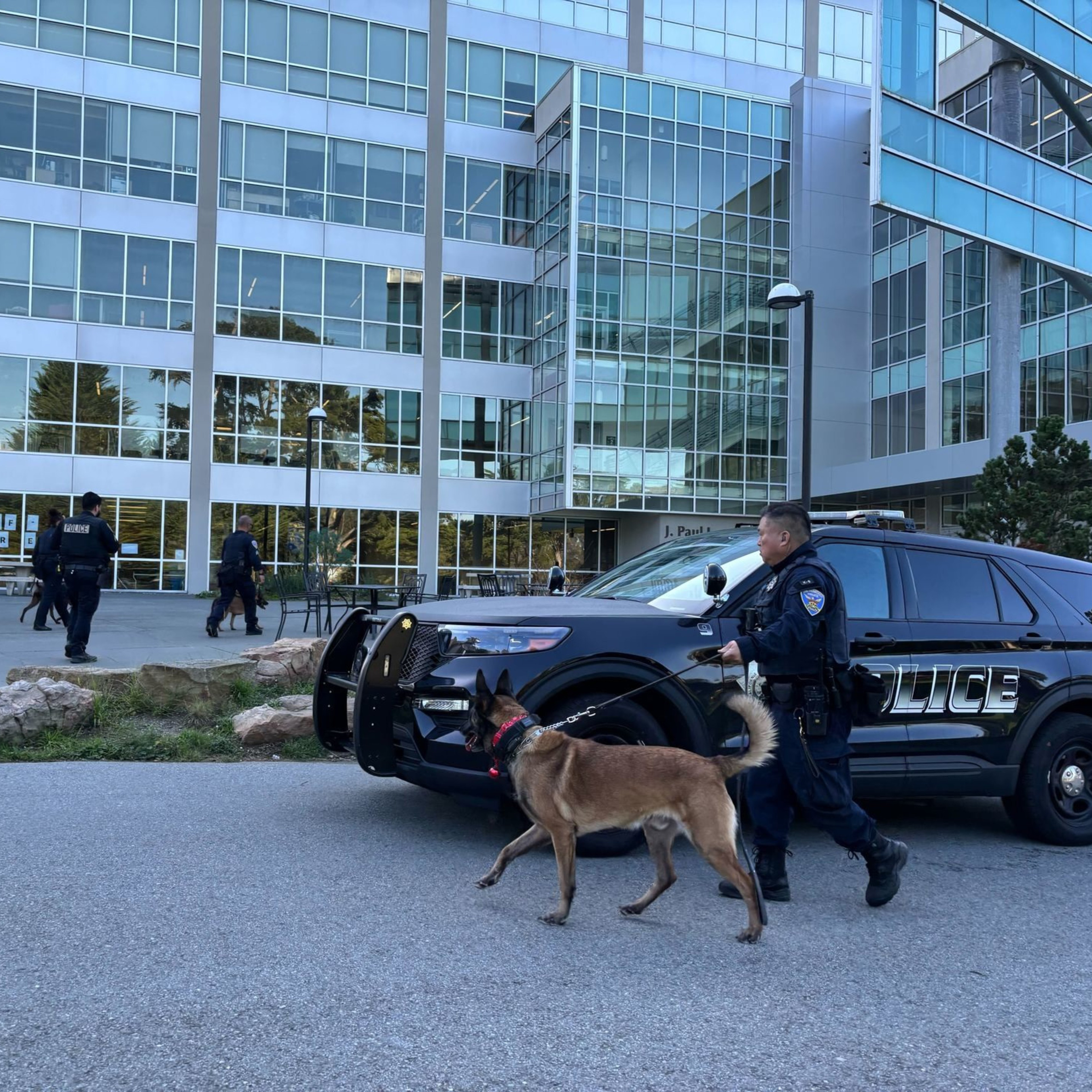 A police officer walks a dog on a leash near a police SUV outside a modern glass building, with two officers in the background.
