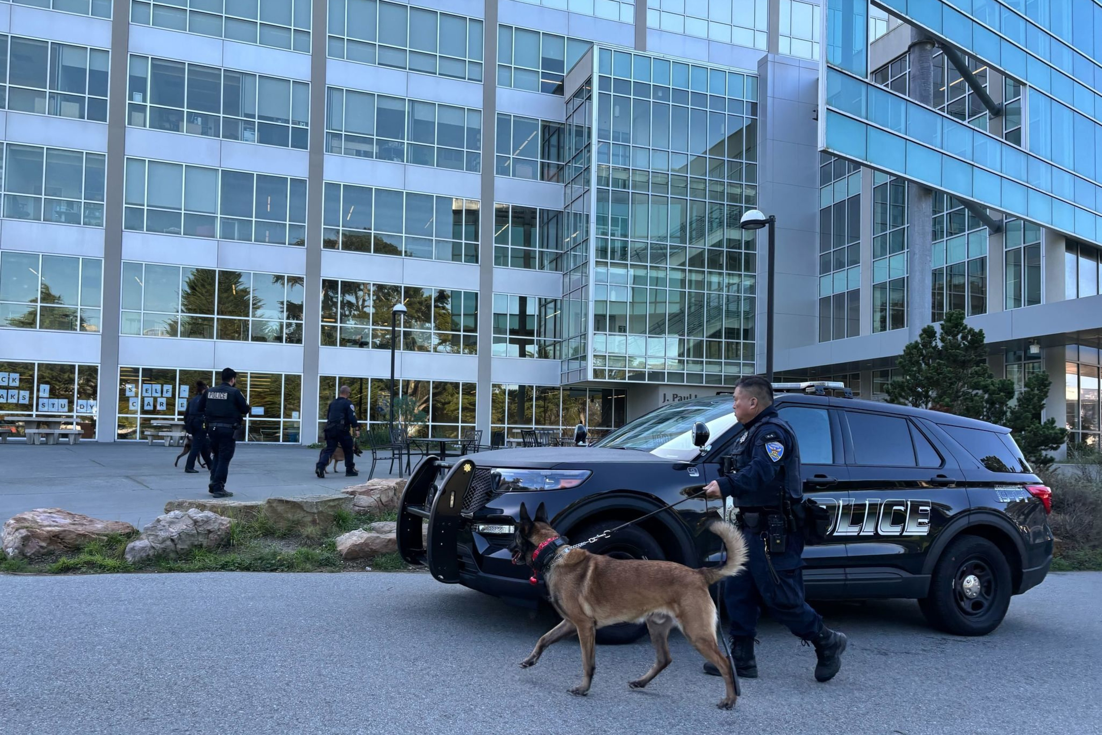 A police officer walks a dog on a leash near a police SUV outside a modern glass building, with two officers in the background.