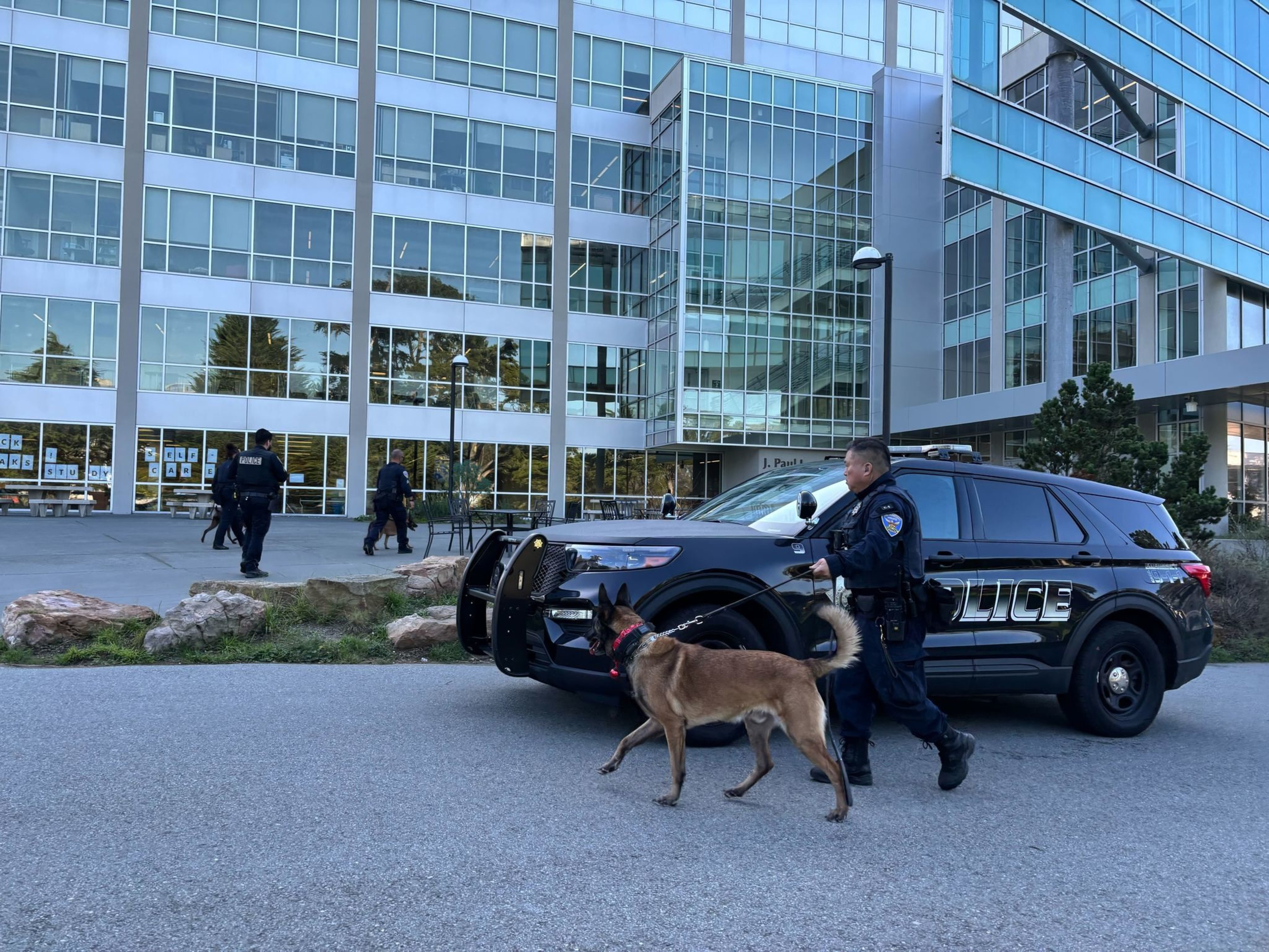 A police officer walks a dog on a leash near a police SUV outside a modern glass building, with two officers in the background.
