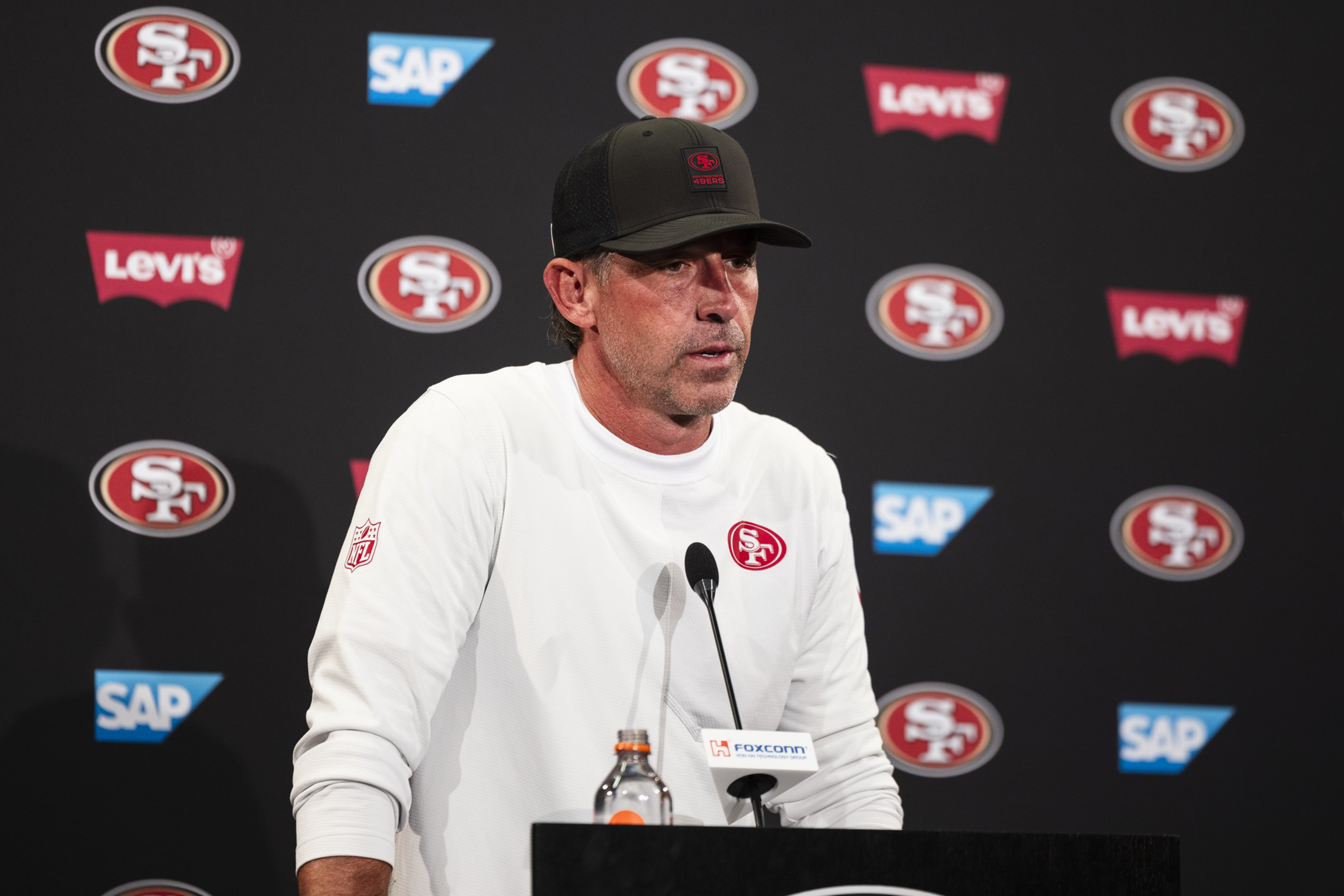 A man in a white long-sleeve shirt and black cap with San Francisco 49ers logos stands at a podium with microphones before a branded backdrop.
