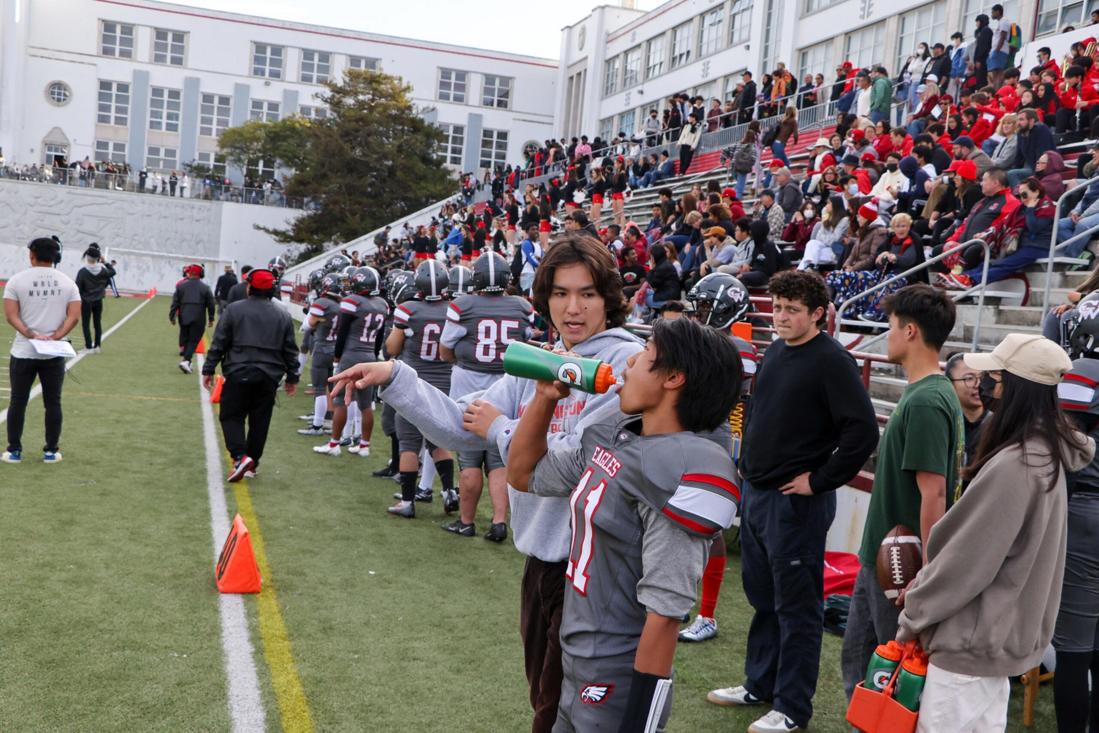 A football player drinks from a green bottle while a coach gestures and talks to him on the sideline, with teammates and spectators in the background.