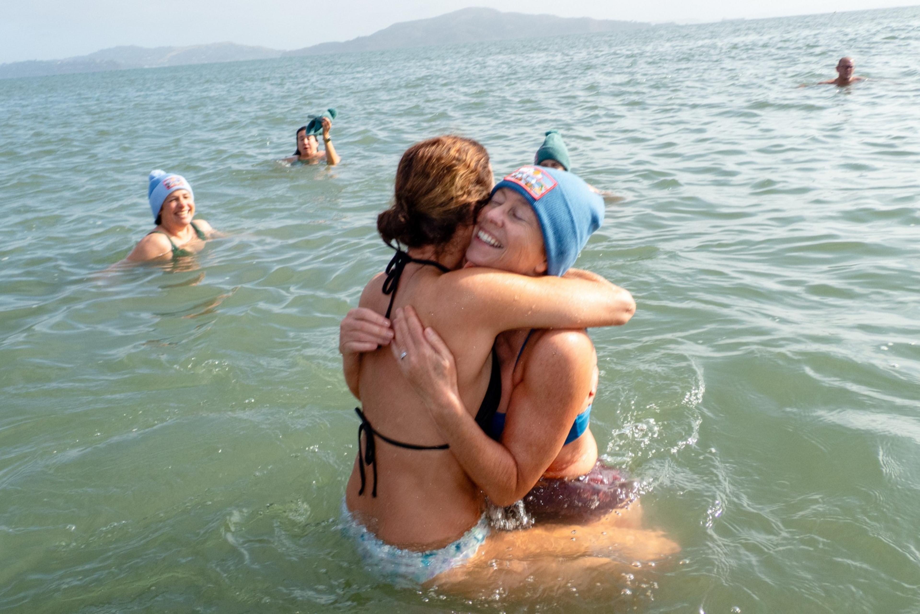 Two people in swimsuits and beanies are joyfully hugging in the water, with three others smiling and swimming nearby under a clear sky.