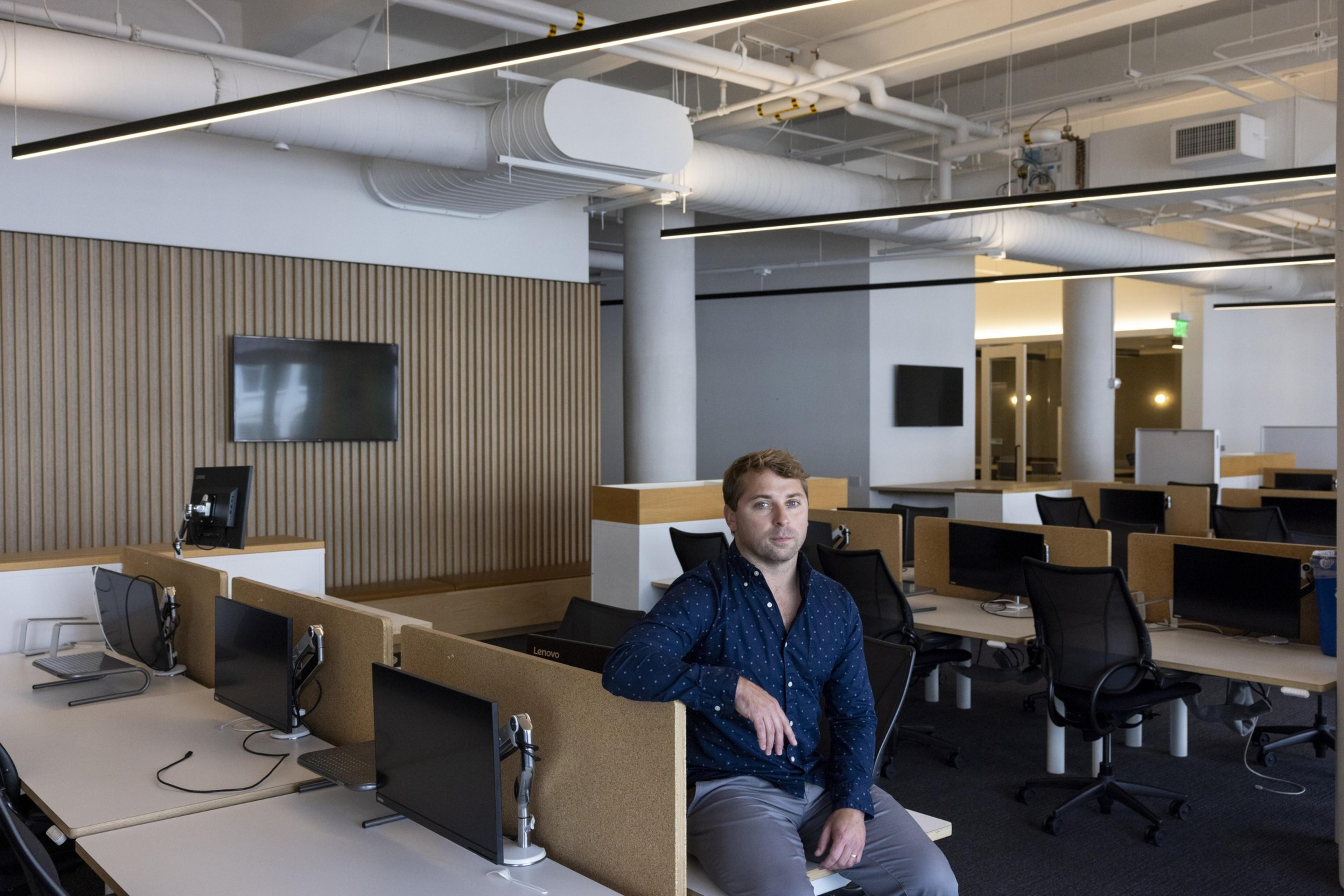 A man is sitting in a modern, open-plan office with multiple empty workstations and monitors, and a wooden accent wall in the background.