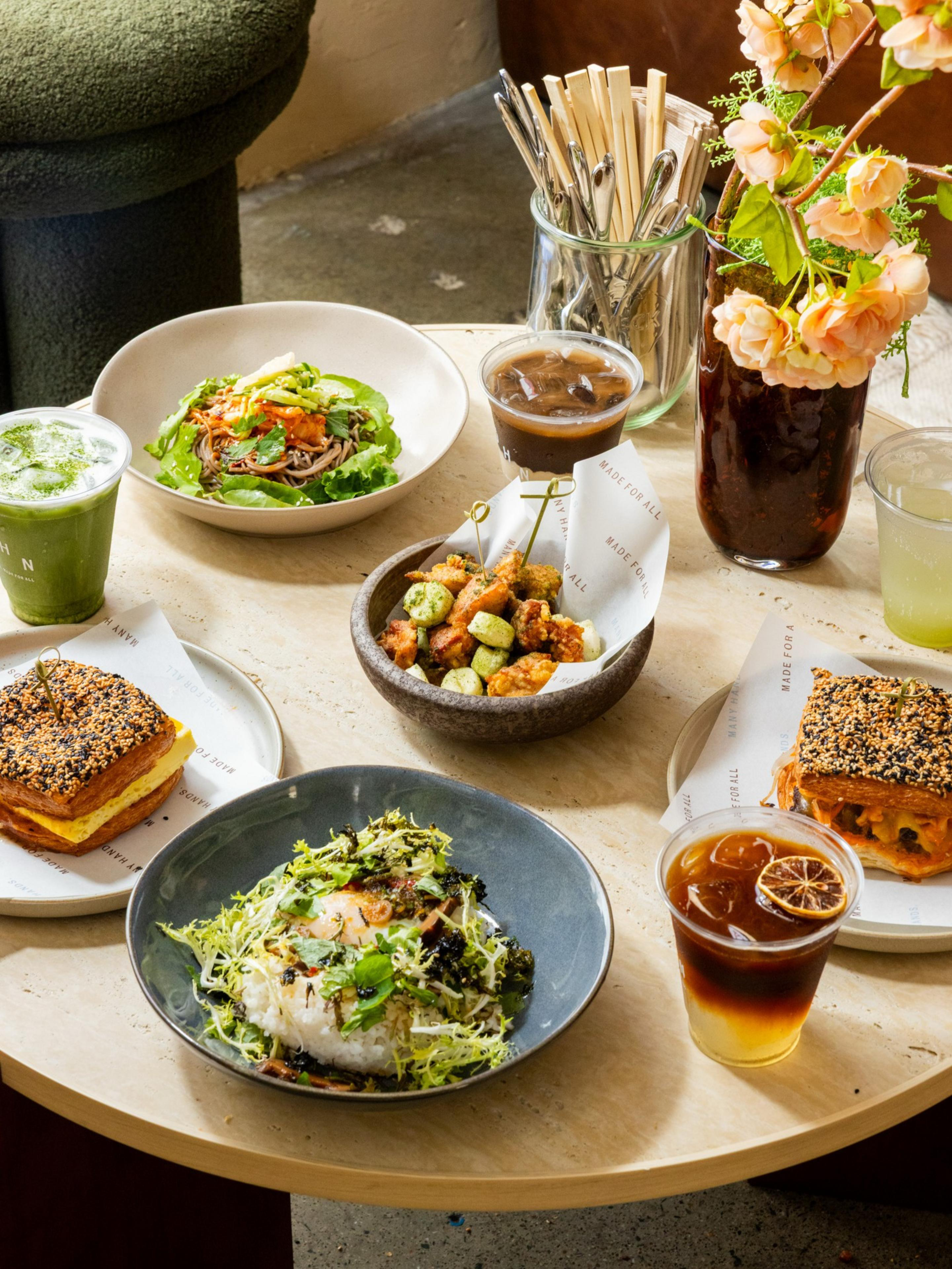 Two sesame-covered sandwiches, a green tea latte, two iced coffees, a dish of fried snacks with edamame, a bowl of salad with mixed greens, and a bowl of noodles or rice with vegetables, all on a round table with chopsticks and a vase of flowers.