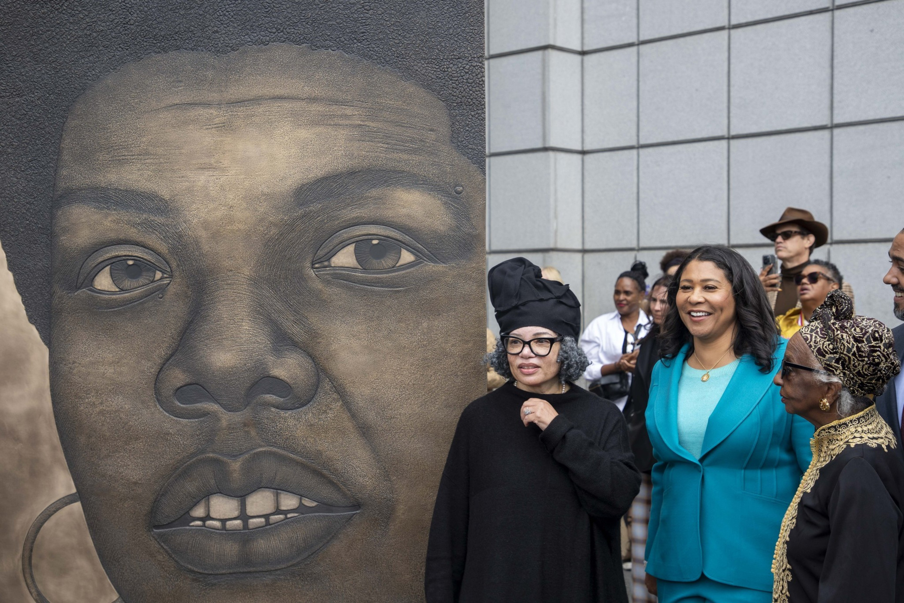 A group of people stand beside a large, detailed mural of a woman's face on a city sidewalk, with one woman in a bright teal outfit smiling.