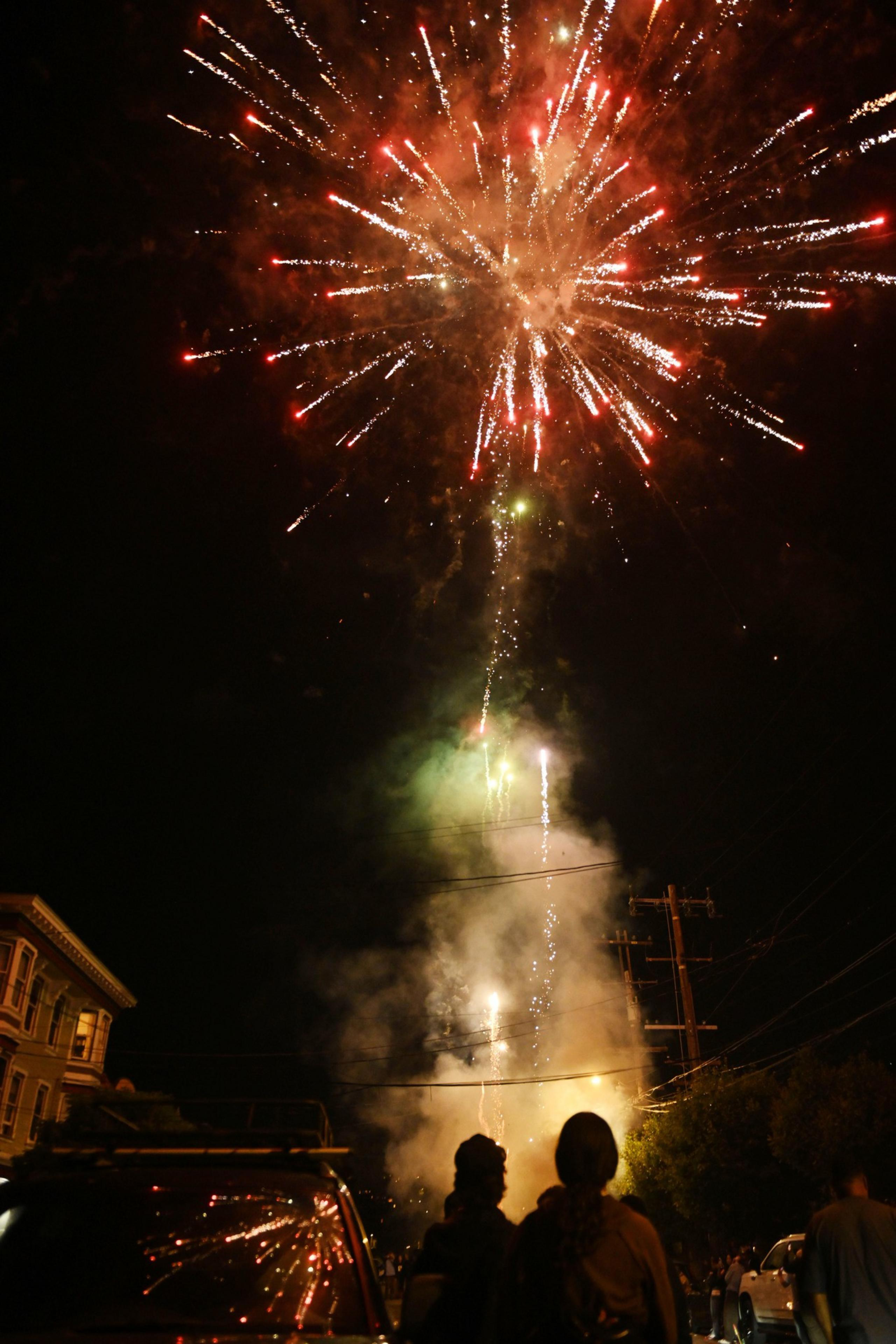 A vivid firework display lights up the night sky with red and white streaks. People watch from a dark street, where cars and buildings are dimly visible amidst the smoke.
