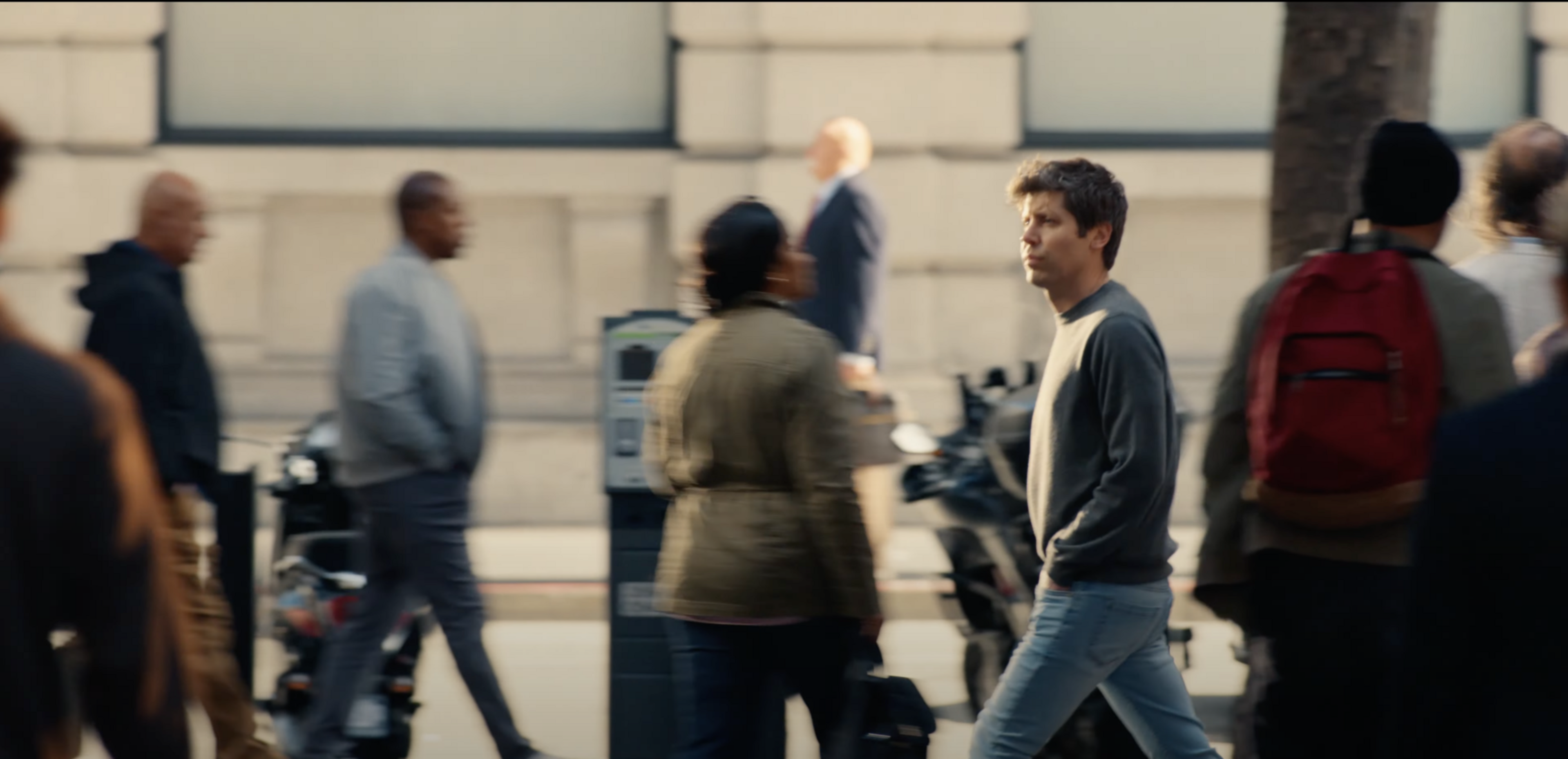 A man in a gray sweater and jeans walks on a city sidewalk. The background shows blurred pedestrians and a building facade, creating a sense of motion.