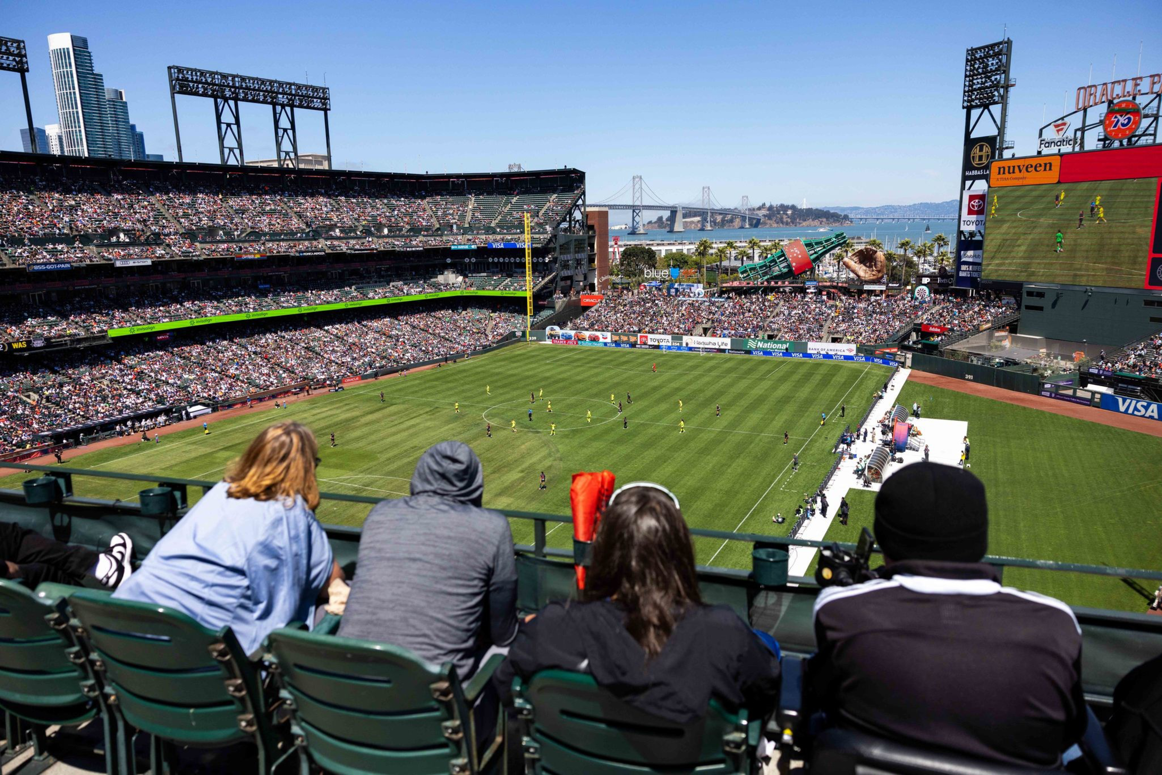 A crowded stadium with fans watching a soccer match on a sunny day, with a large screen displaying the game and a cityscape, including a bridge, in the background.