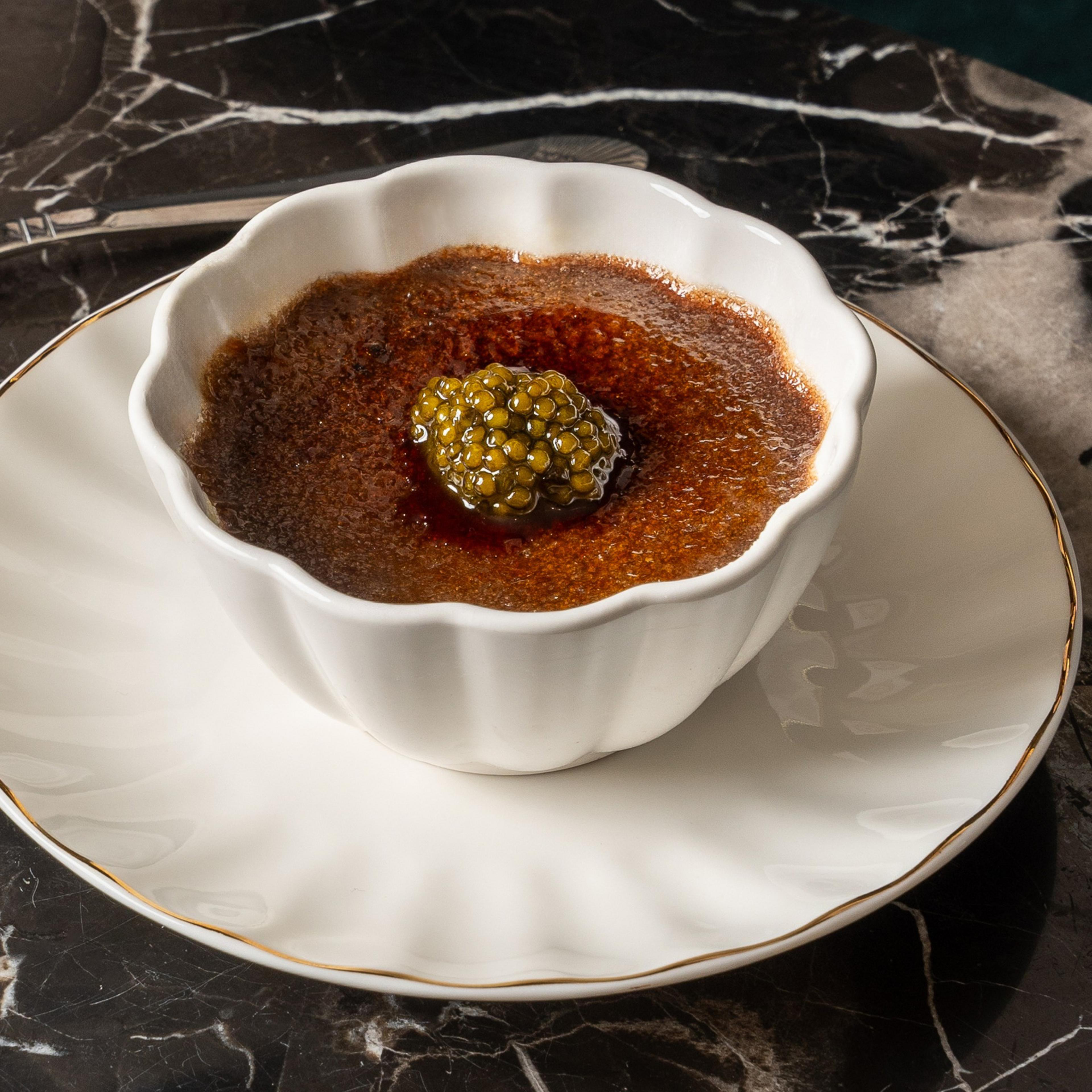 A white fluted bowl holds crème brûlée topped with caviar, sitting on a matching plate with a gold rim, placed on a dark, marbled surface.